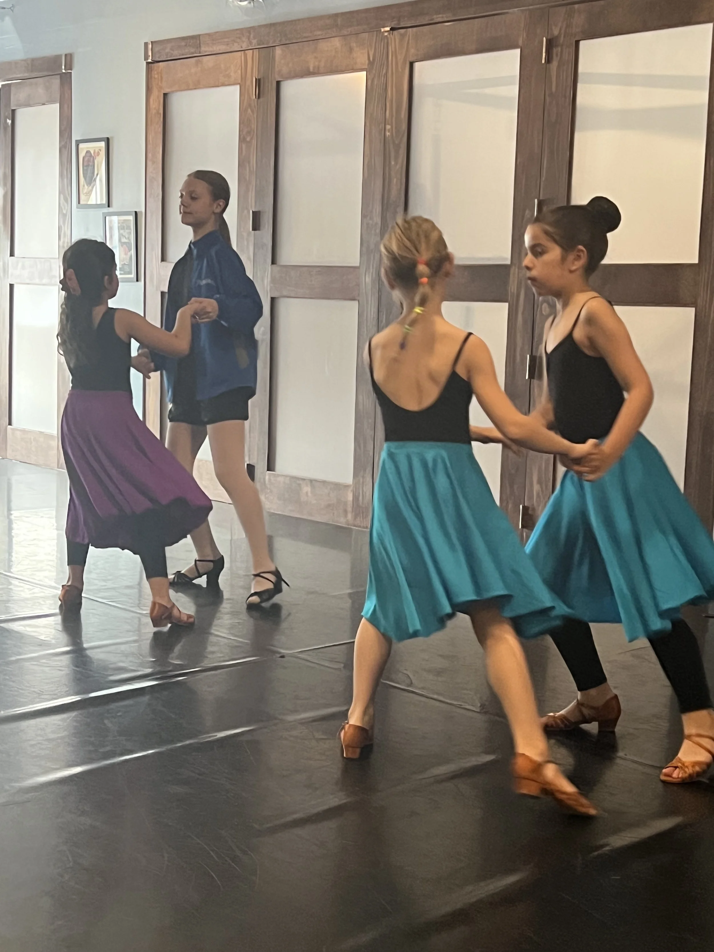 Four young girls practicing a dance in a studio with wooden wainscoting and framed art on the wall.