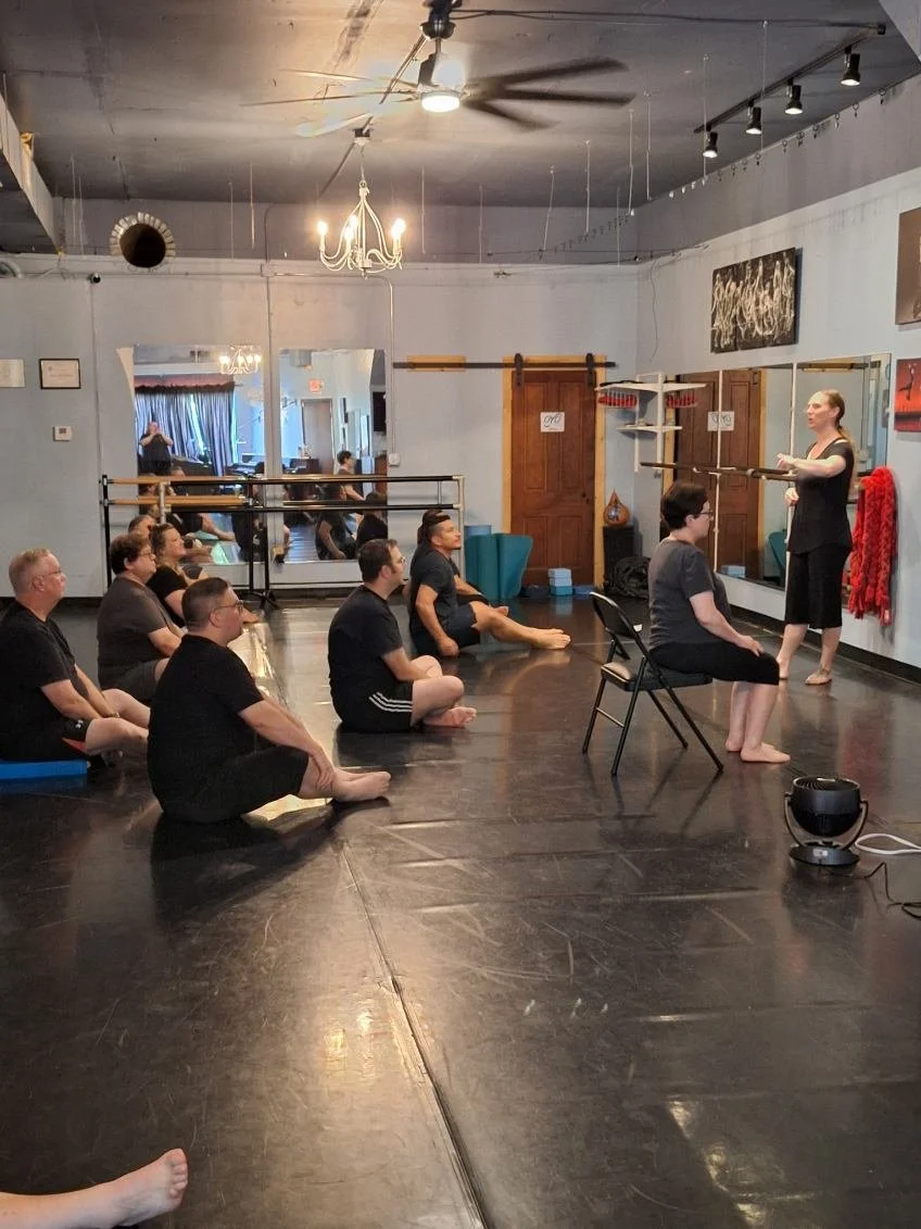 A yoga or meditation class with participants sitting on the floor facing a teacher who is standing and holding a wooden stick. The room has a dark floor, mirrors on the walls, and various decorations and artwork. Some students are seated on cushions, and a few are sitting on chairs. The instructor appears to be demonstrating or explaining something to the class.