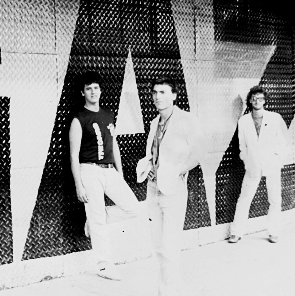 Three young people standing in front of a decorative wall with a large star design. The person on the left wears a sleeveless shirt and white pants. The person in the middle wears a light-colored jacket and pants, with his hands in his pockets. The person on the right wears glasses, a white jacket, and white pants.