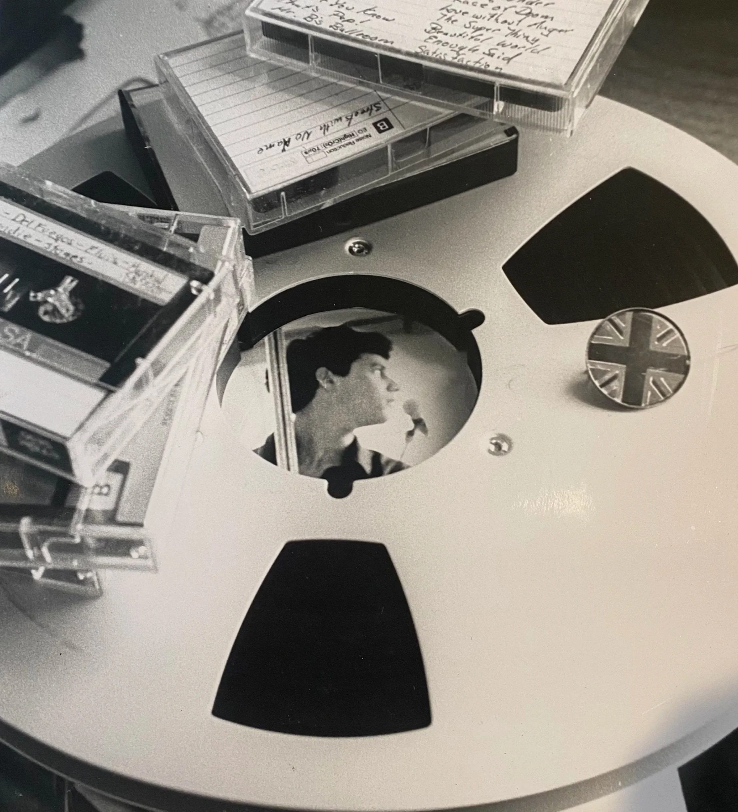 A black-and-white view of a person’s face in a circular photo frame on a music reel-to-reel tape recorder, surrounded by cassette tapes and handwritten notes.