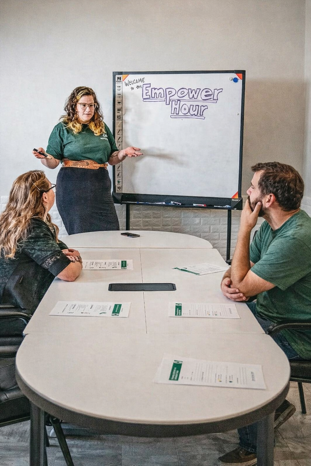 A woman giving a presentation to two seated individuals in a meeting room with a whiteboard that reads "Empower Hour" and a welcome message.