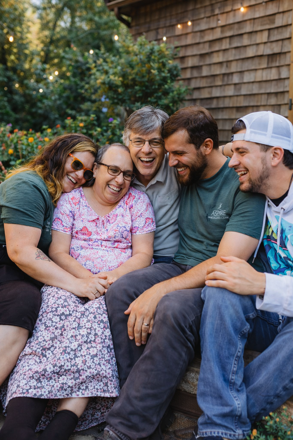 A group of five people, three women and two men, smile and laugh together outdoors in a garden, sitting close and showing a joyful moment.