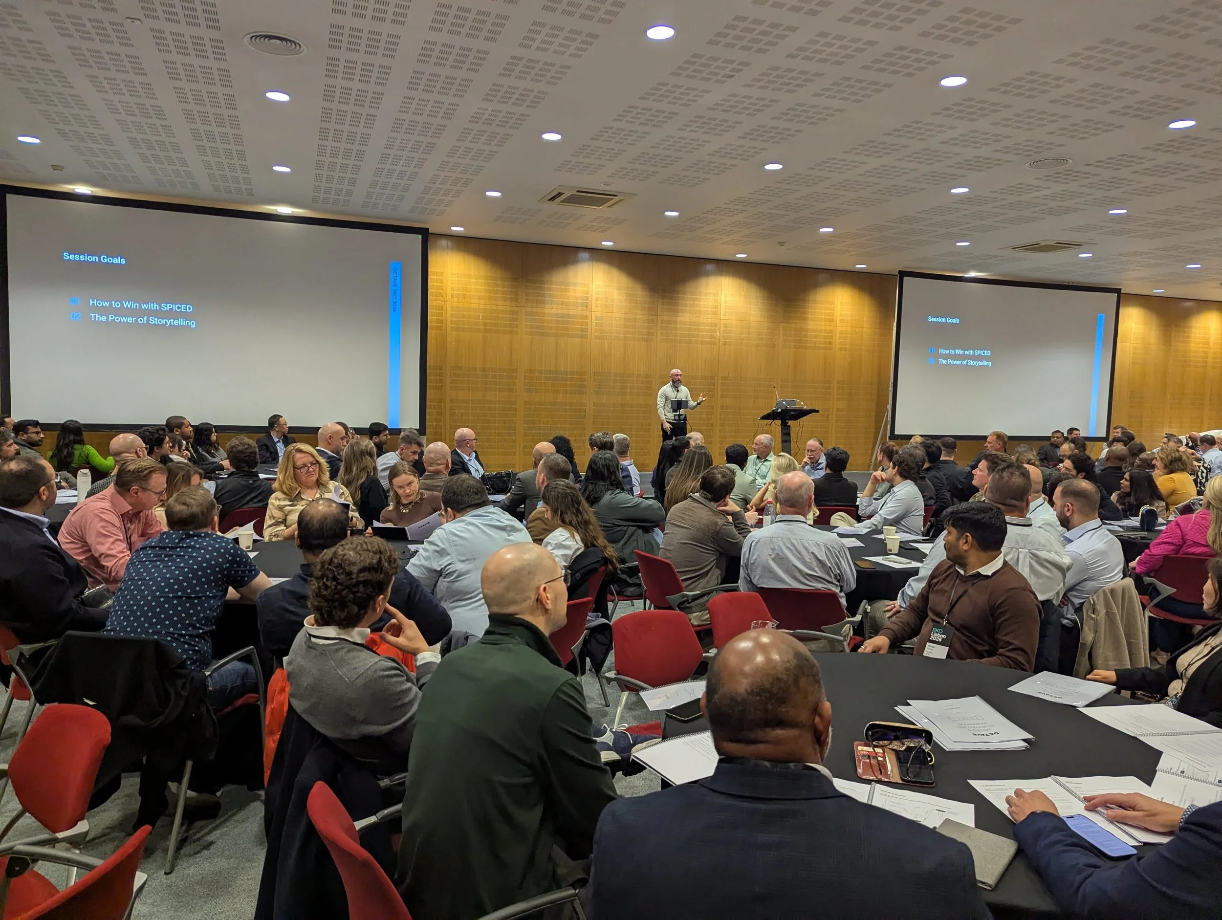 A large conference room filled with attendees seated at round tables, listening to a speaker at a podium. Two large screens display presentation slides with session goals.