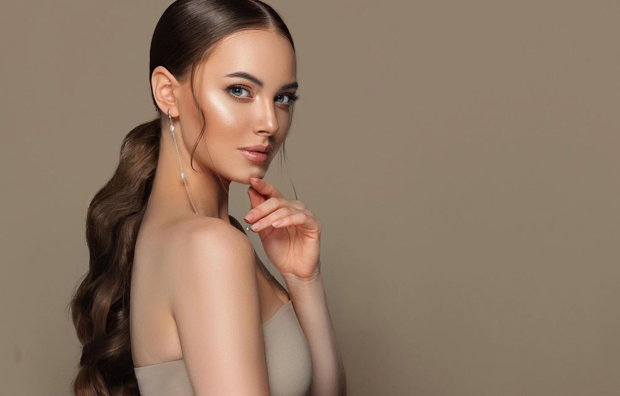 A woman with long brown hair, wearing makeup, a strapless beige top, and dangling earrings, posing against a plain beige background.