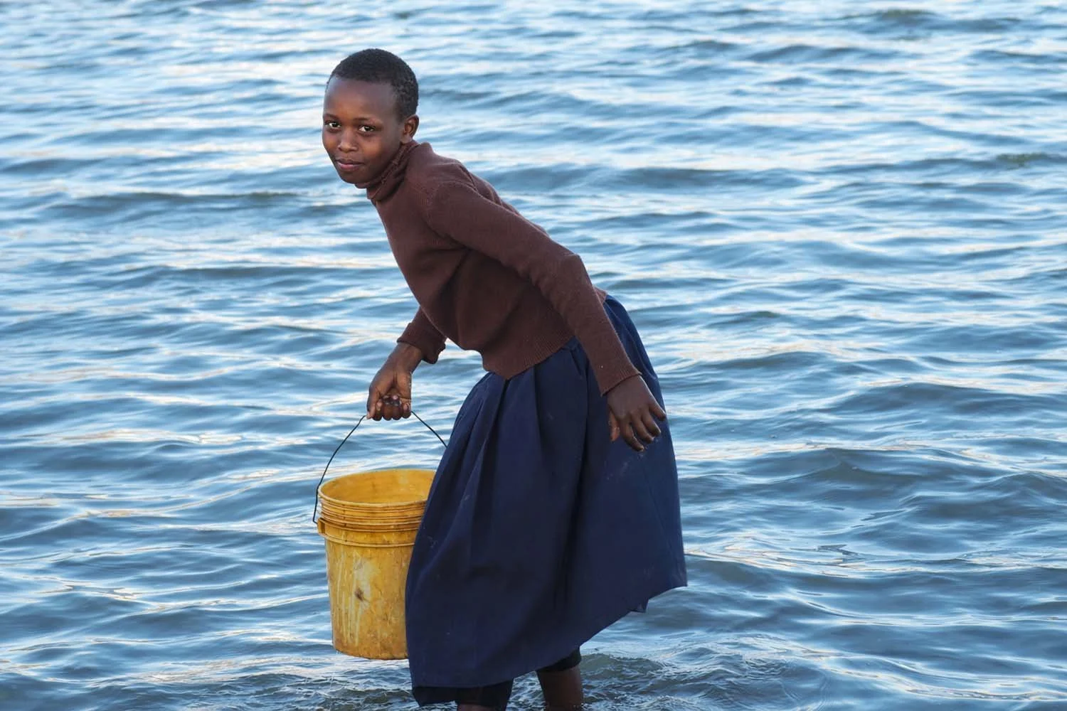 A young girl standing in water, holding a yellow bucket.