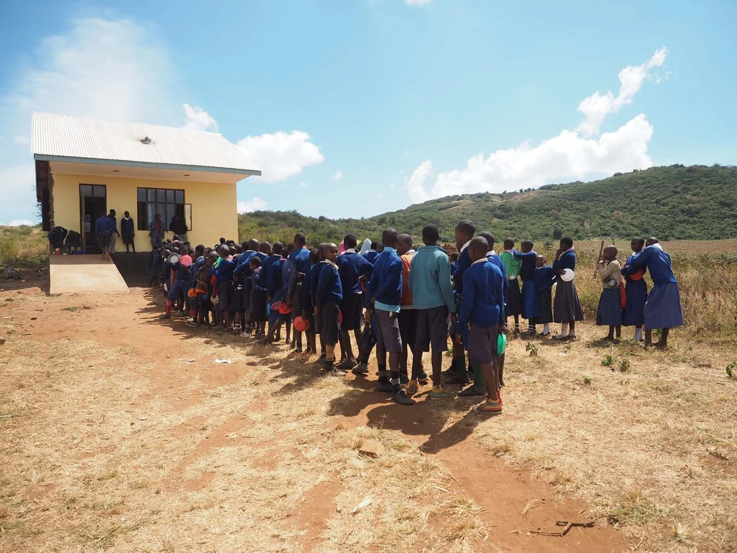 A group of school children waiting in line outside a small yellow school building on a sunny day, with hills and blue sky in the background.