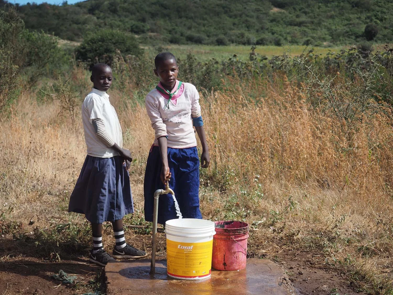 Two schoolgirls in uniform and stripes socks are collecting water from a tap in a rural landscape with dry grass and hills in the background.