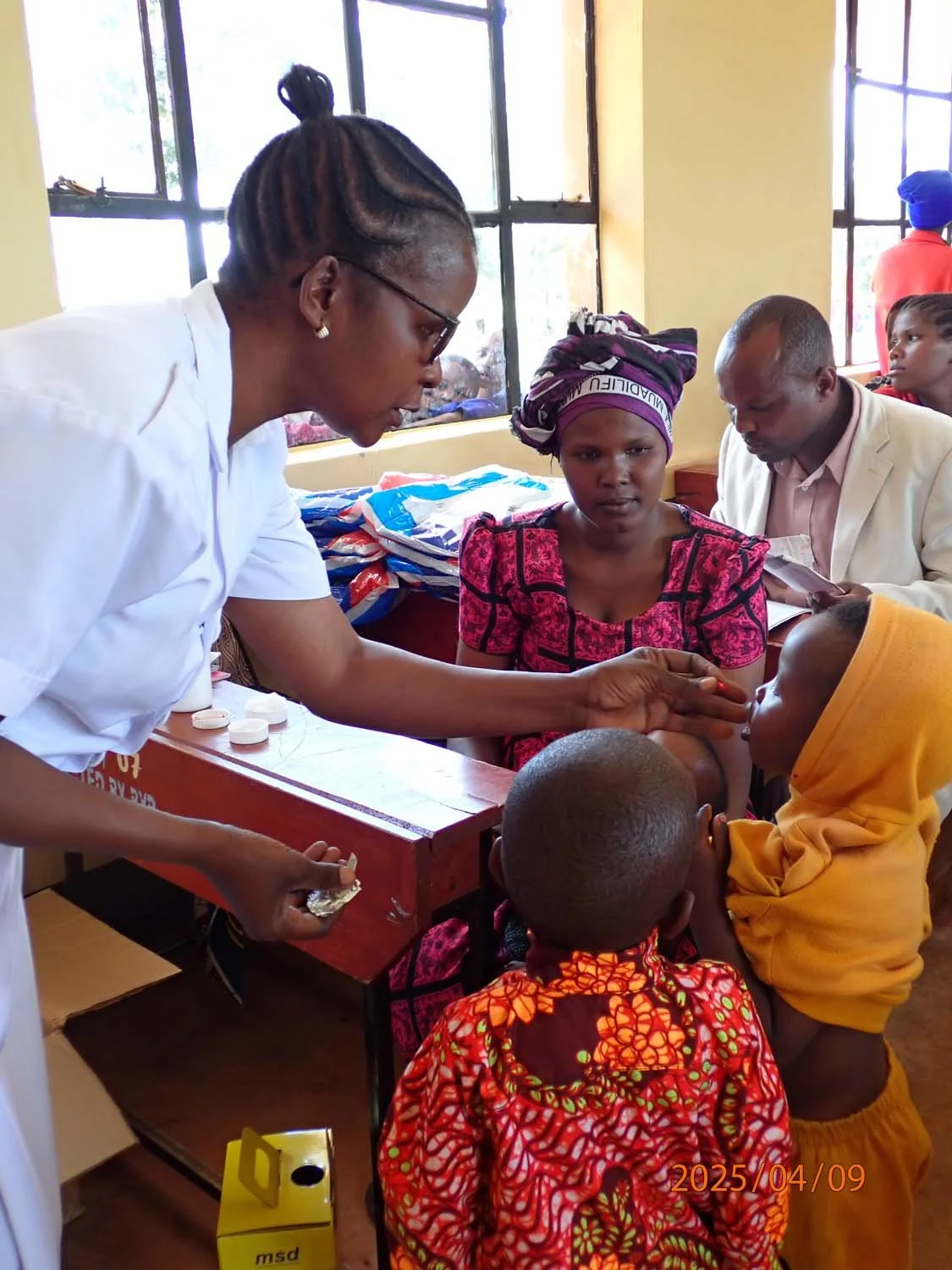 A healthcare worker administering medicine to a young girl at a clinic, with children and adults waiting and seated around. The scene is indoors with large windows, and there are supplies and medication on the table.