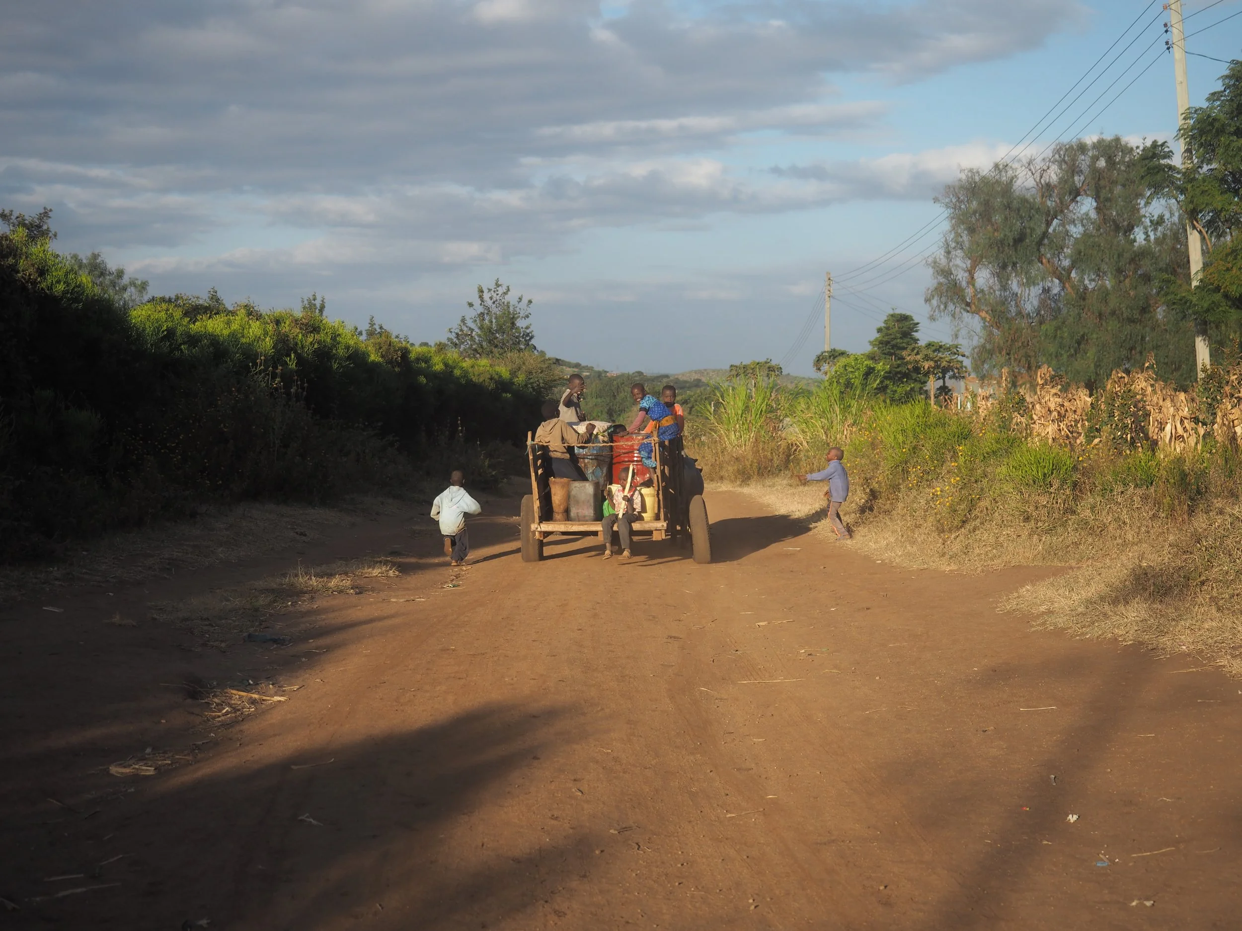 Children and a few people riding and walking near a parked tractor on a dirt road with greenery and power lines in the background.