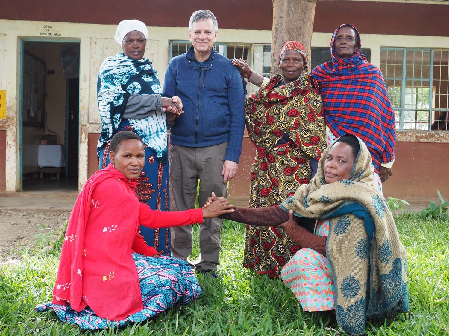 A group of six people, five women and one man, outdoors in front of a building, with two women kneeling and shaking hands, the others standing around them, all dressed in colorful clothing and traditional fabrics.