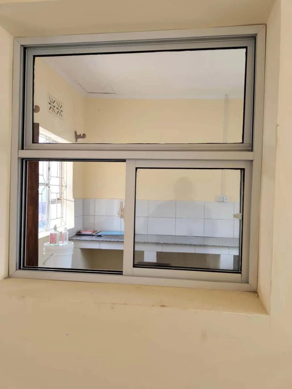 View through a window showing a kitchen with a countertop, window grill, white tiles, and some items on the counter.