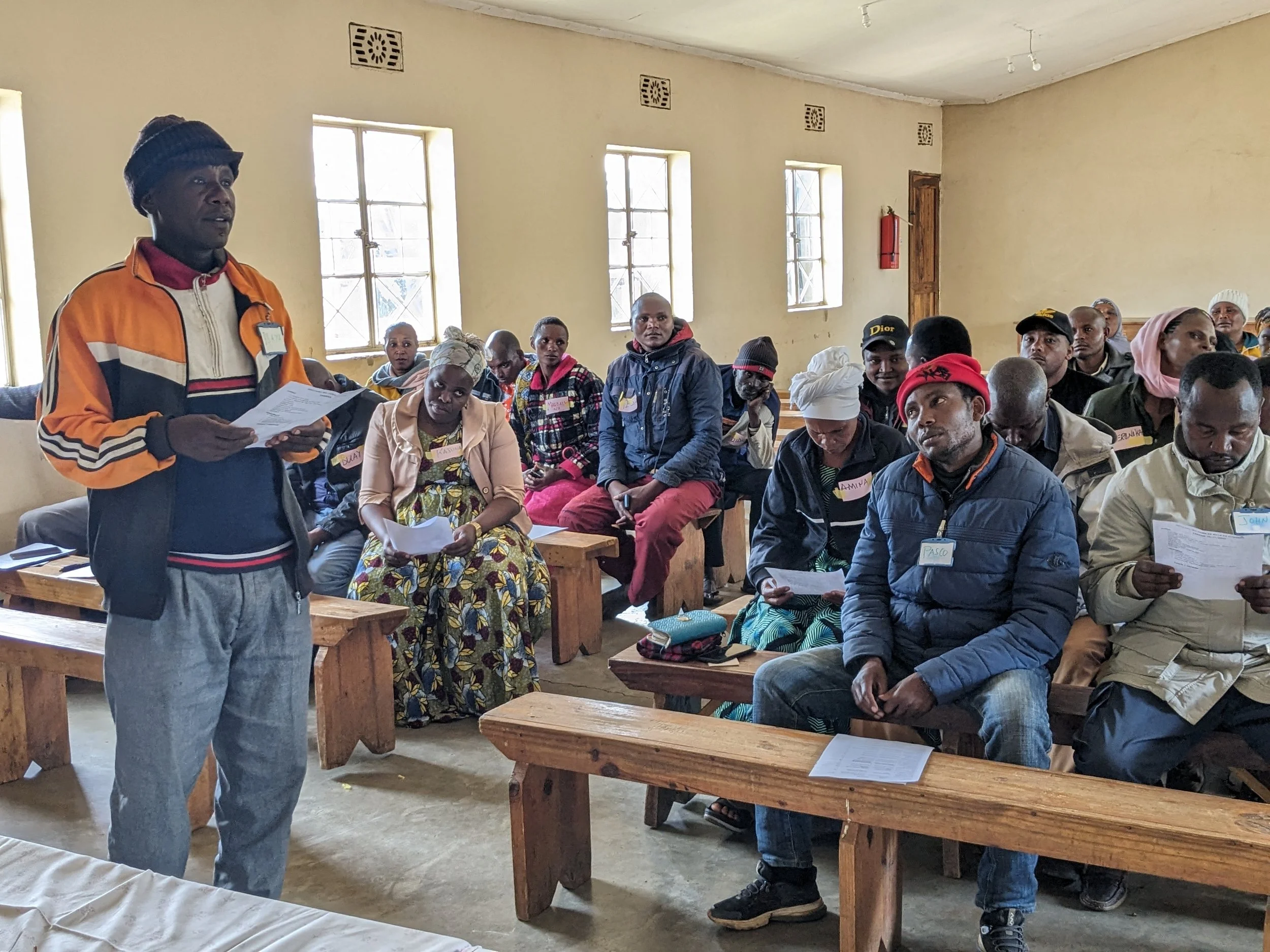 A man standing and speaking to an audience in a room with beige walls and multiple windows, while holding papers. The audience, some taking notes, is seated on wooden benches.