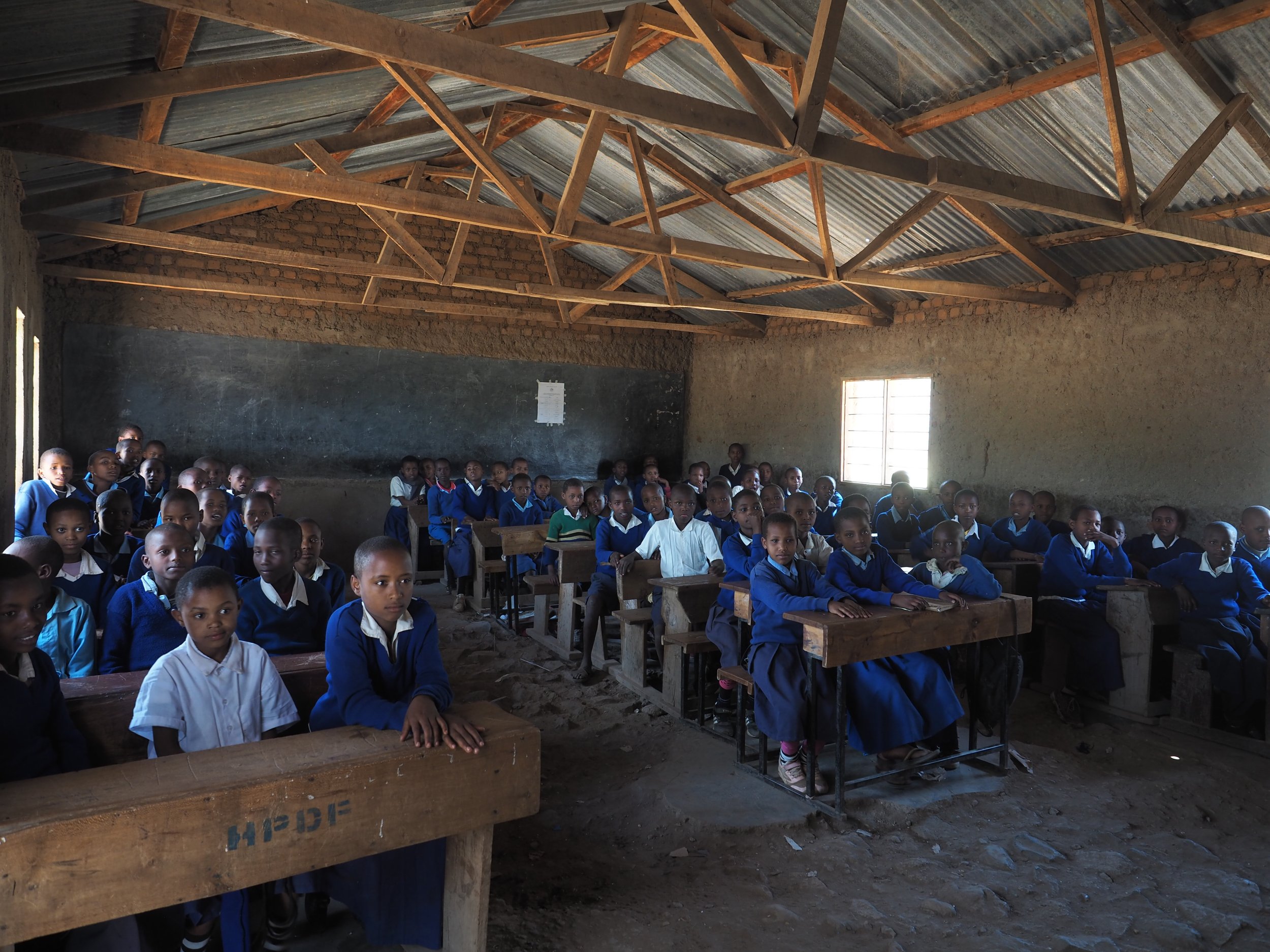 A classroom with students sitting at wooden desks, some looking at the camera, inside a rustic building with a tin roof and unplastered brick walls.