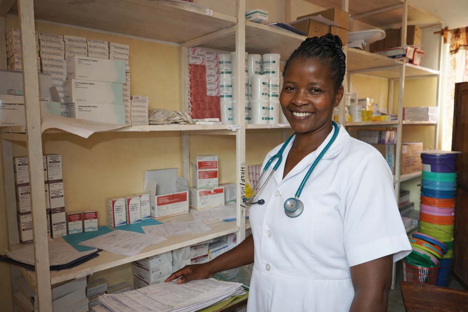 A smiling female healthcare worker with a stethoscope around her neck standing in a pharmacy or medical storage room filled with medication boxes and supplies.