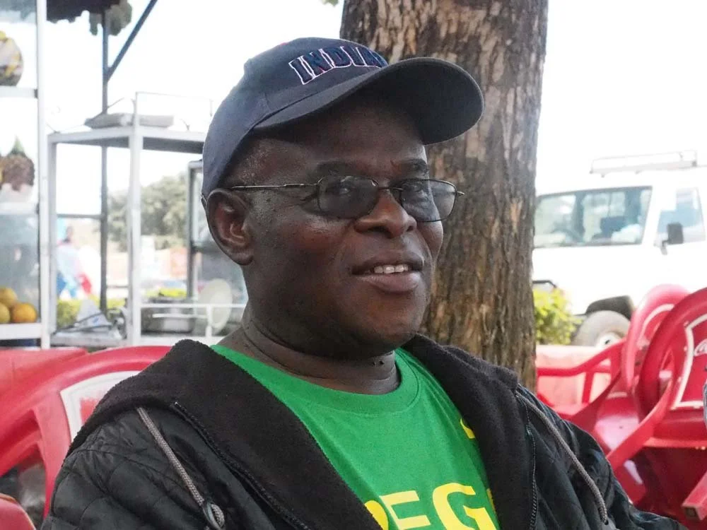 A man wearing a baseball cap, glasses, a green shirt, and a black jacket sitting outdoors near a tree, with red plastic chairs and a vehicle in the background.