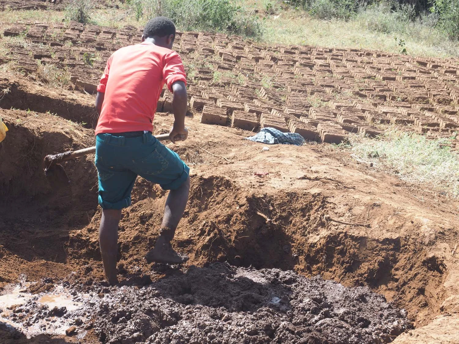 A young man working outdoors, digging or shoveling dirt on a hillside with layered bricks in the background.