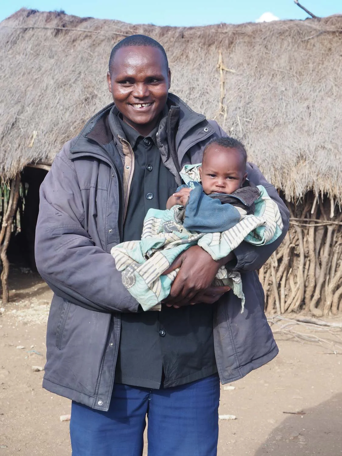 A man smiling and holding a young child wrapped in a blanket standing outdoors in front of traditional thatched-roof huts.