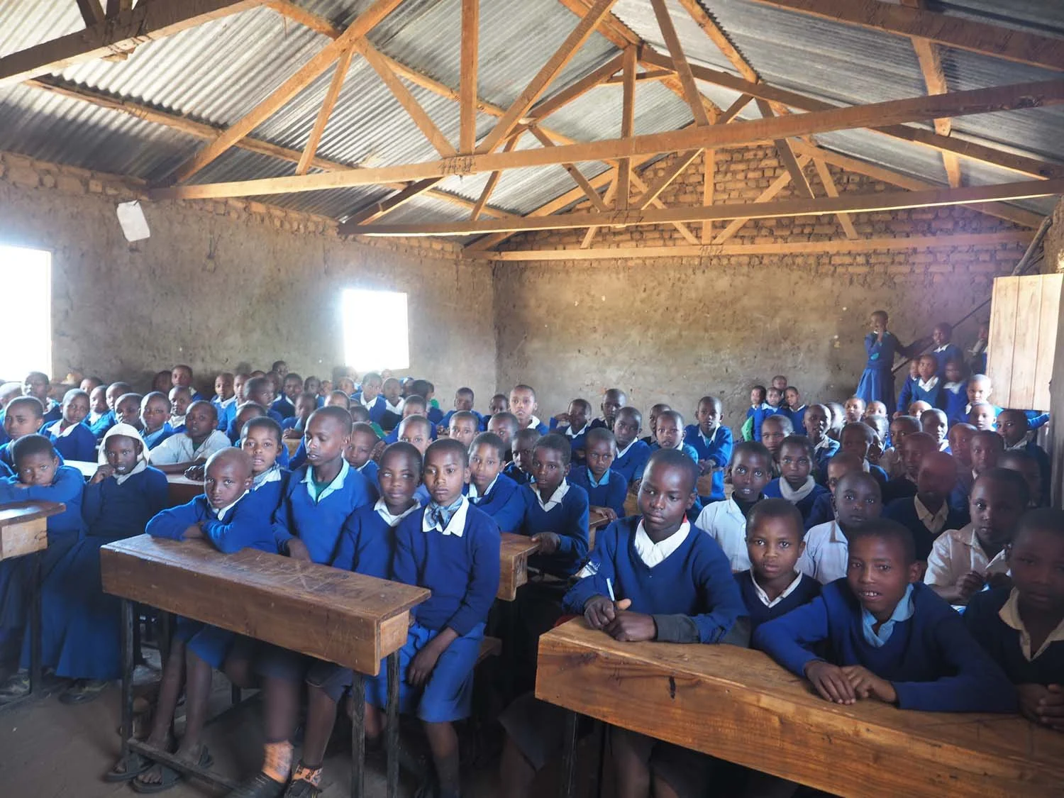A classroom full of young students wearing blue uniforms, seated at wooden desks inside a simple building with exposed brick walls and a corrugated metal roof.