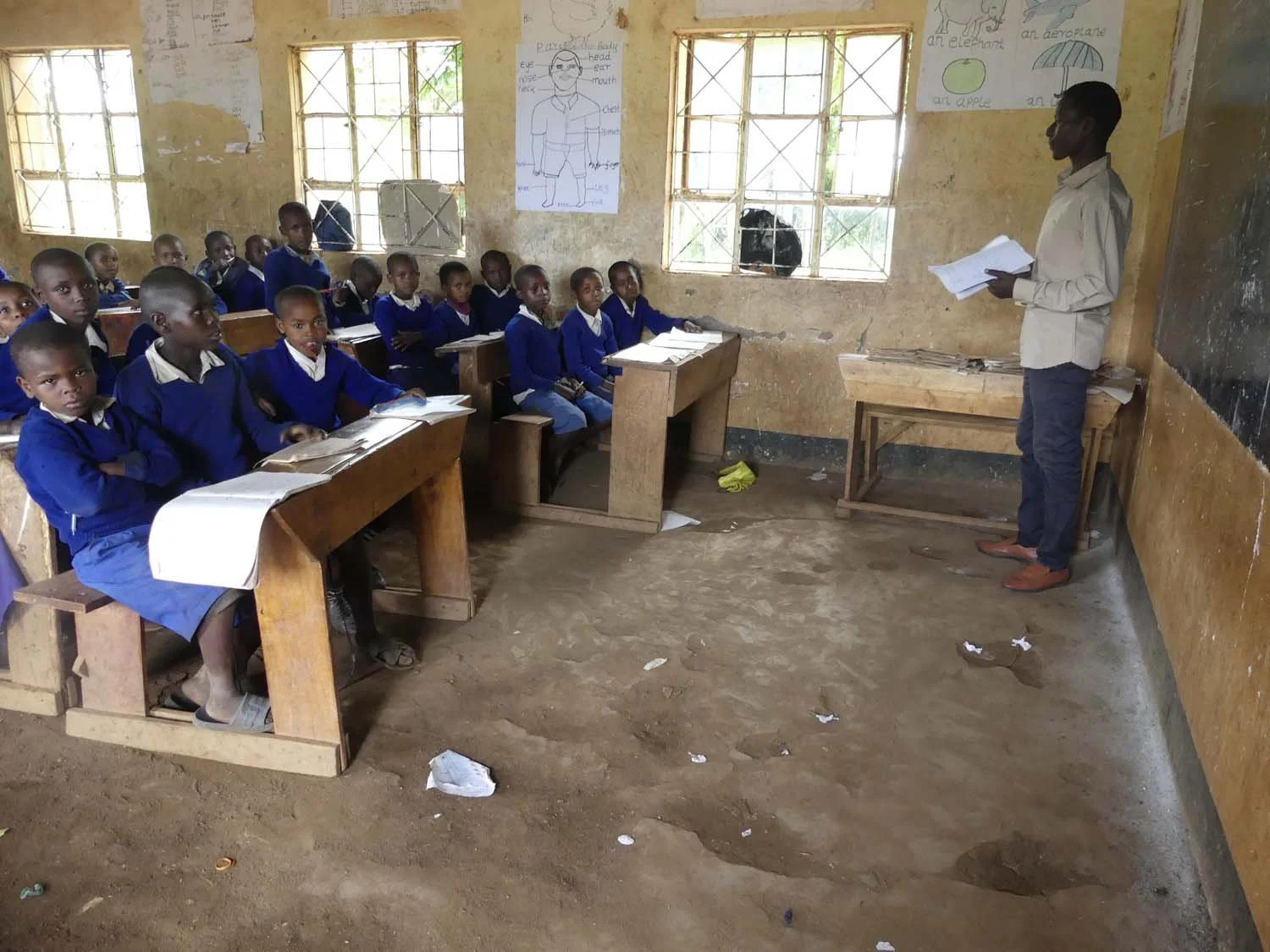 A classroom with young students in blue uniforms sitting at wooden desks, listening to a teacher standing at the front with notes. The classroom has bare, worn walls and dirt floor, and educational posters on the walls.