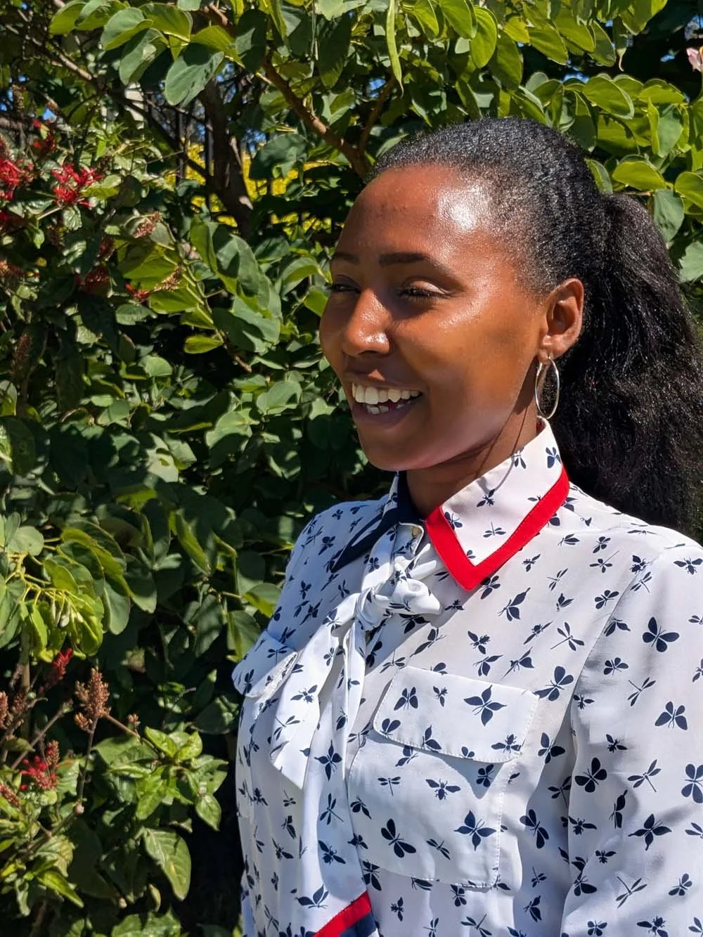 A woman smiling outdoors among green foliage, wearing a white blouse with a blue floral pattern, red accents, hoop earrings, and her hair pulled back.