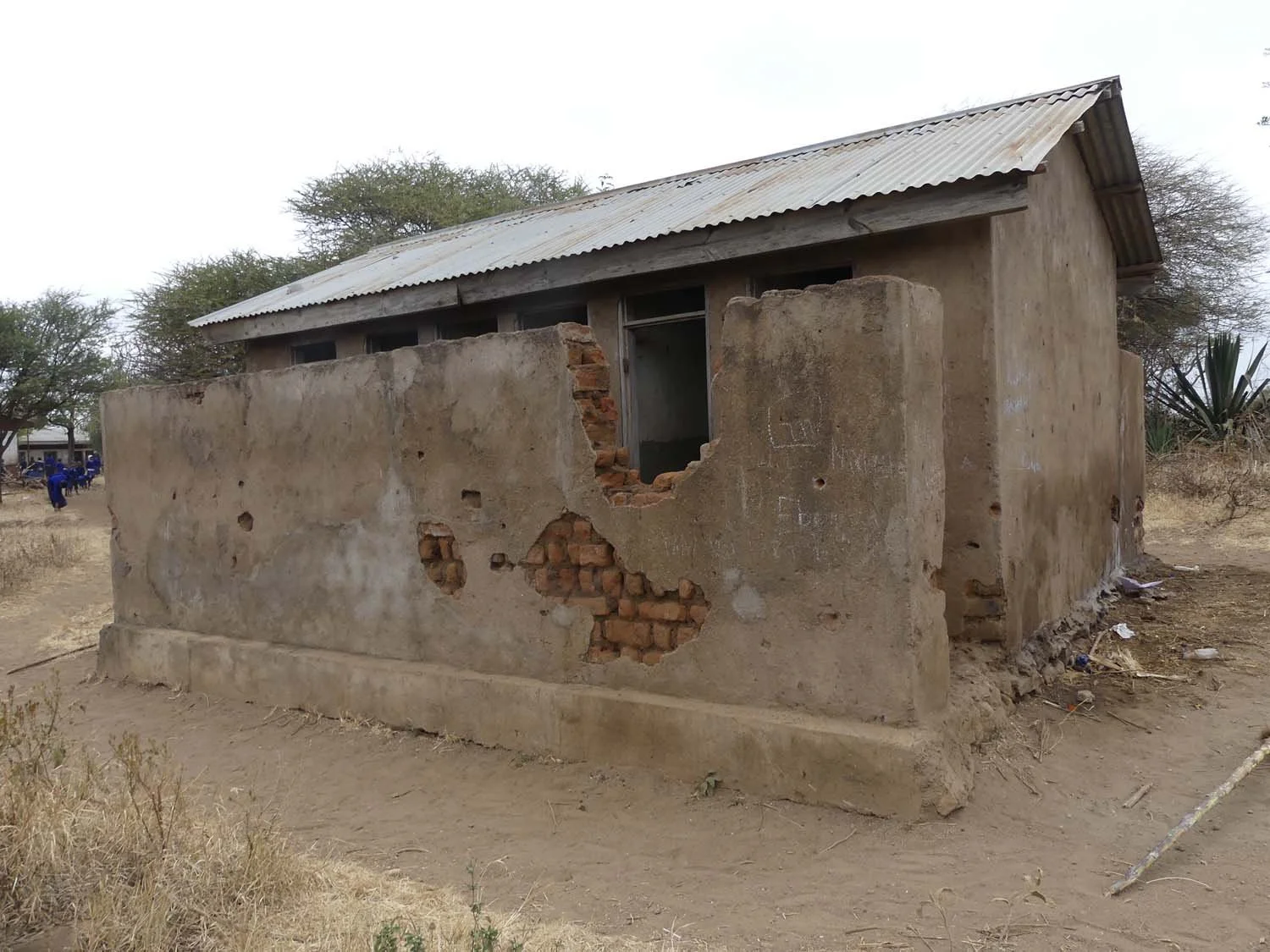 A small, partially damaged building with brick and concrete walls, a metal roof, and no windows, in a dry, arid environment.