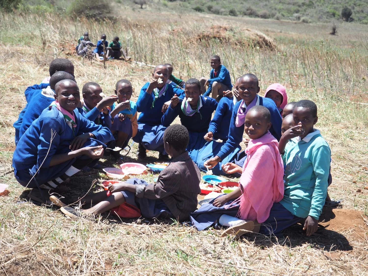 Group of children sitting on dry grass in a rural landscape, eating food from bowls.