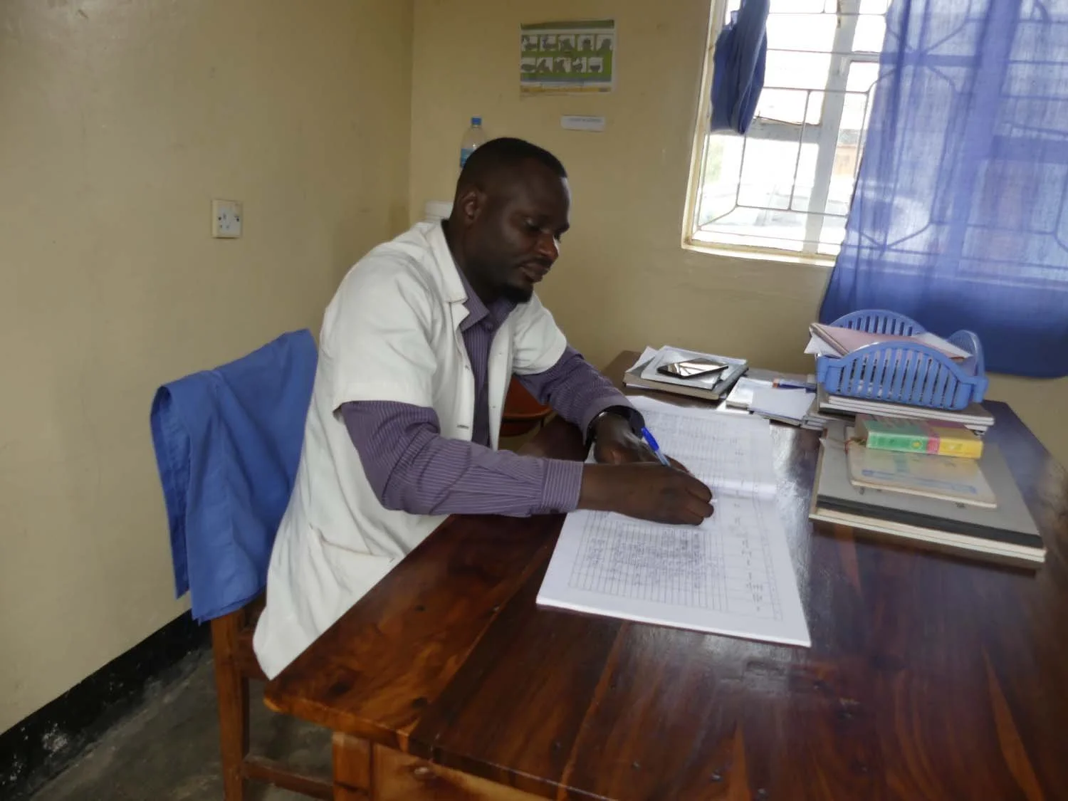 A man seated at a wooden desk writing in a large ledger. The desk has stacks of papers, folders, and a blue file organizer. Behind him, there are windows with blue curtains and a beige wall with a small poster or chart.
