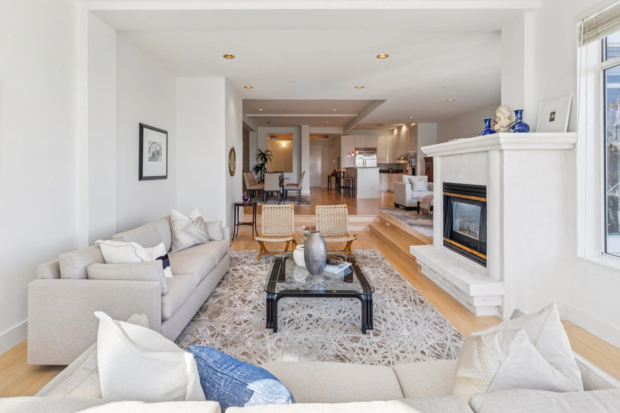 Interior view of a modern living room with white walls, beige sofas, a glass coffee table, a fireplace, and a large window.