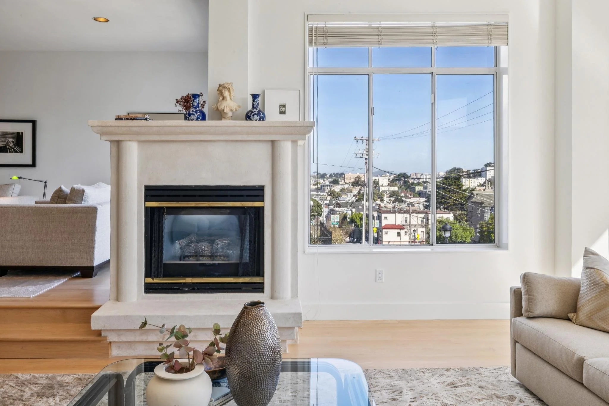 Living room with a fireplace, large window showing a view of a city with hills, beige sofa, decorative vases on the mantle, and a coffee table with a vase and a potted plant.