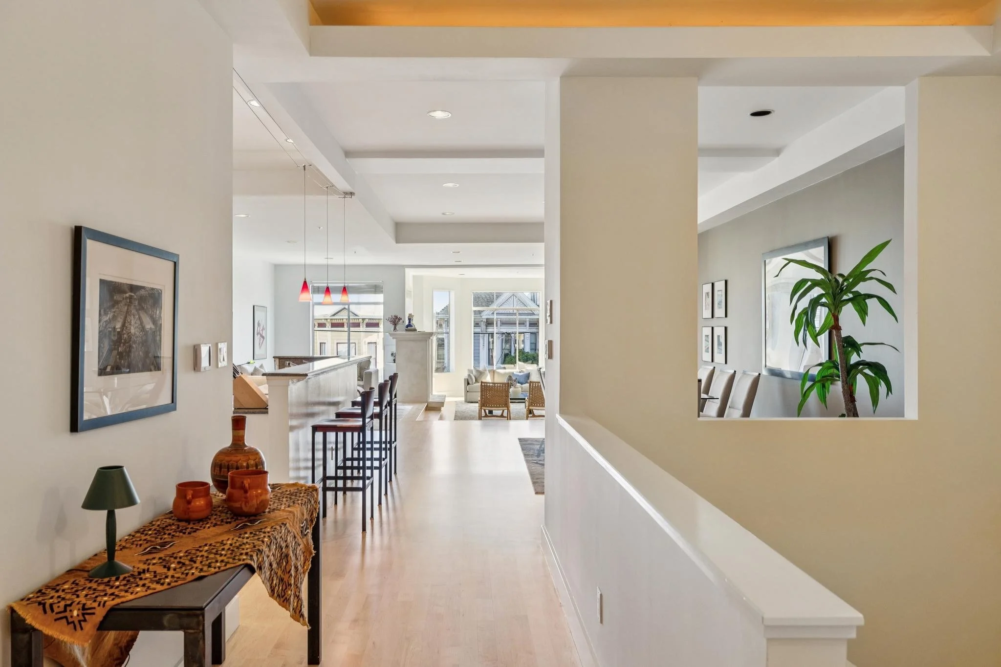 Interior view of a modern home with a hallway leading to a living area, featuring a high ceiling, large windows, wall art, a table with decorative pottery, and a tall potted plant.