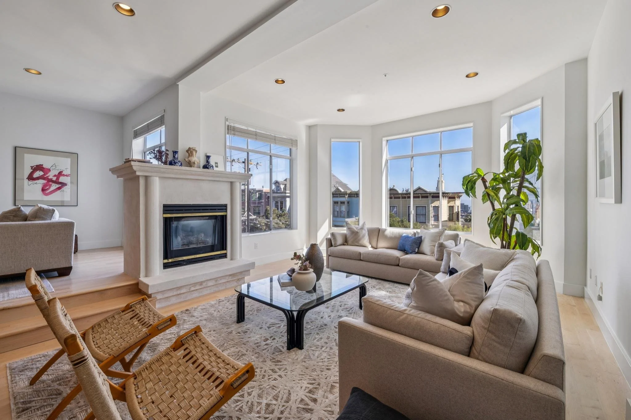 Bright living room with white walls, large bay windows, beige sofas, a glass coffee table with vases, a patterned rug, and a tall potted plant near the window.