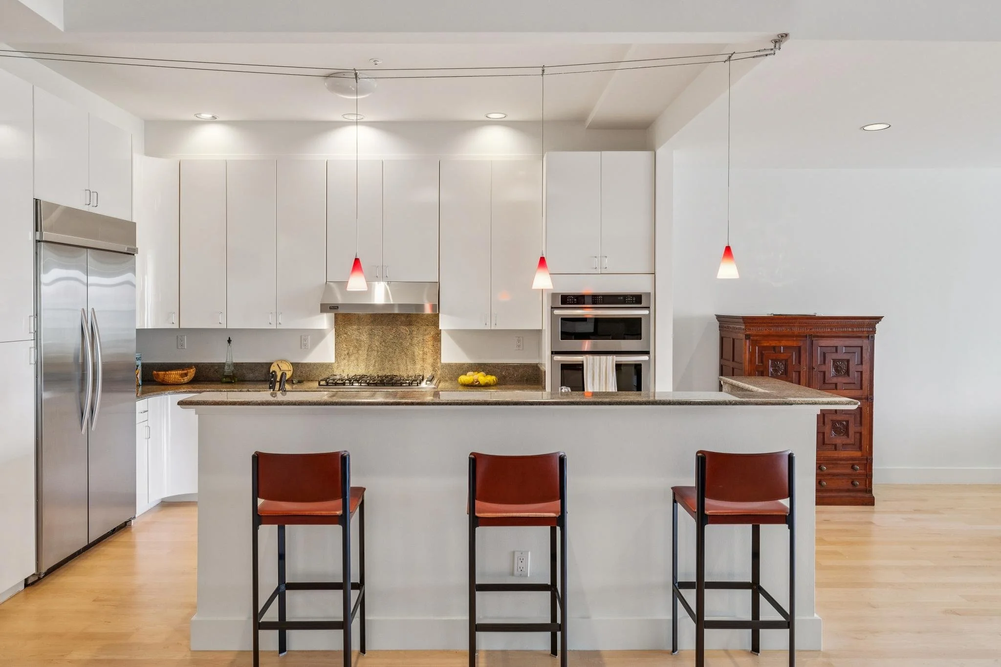 Modern kitchen with white cabinets, stainless steel appliances, granite countertops, three red pendant lights above a breakfast bar, and three red bar stools in the foreground.