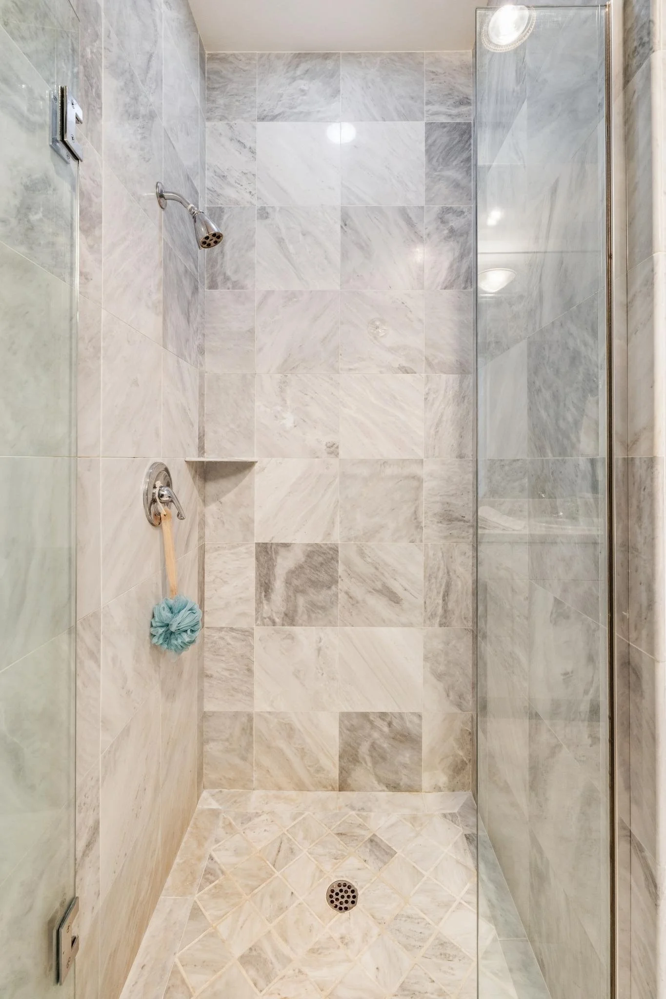 Empty walk-in shower with marble tiles, a showerhead, and a glass door, with a teal shower pouf hanging on the wall.