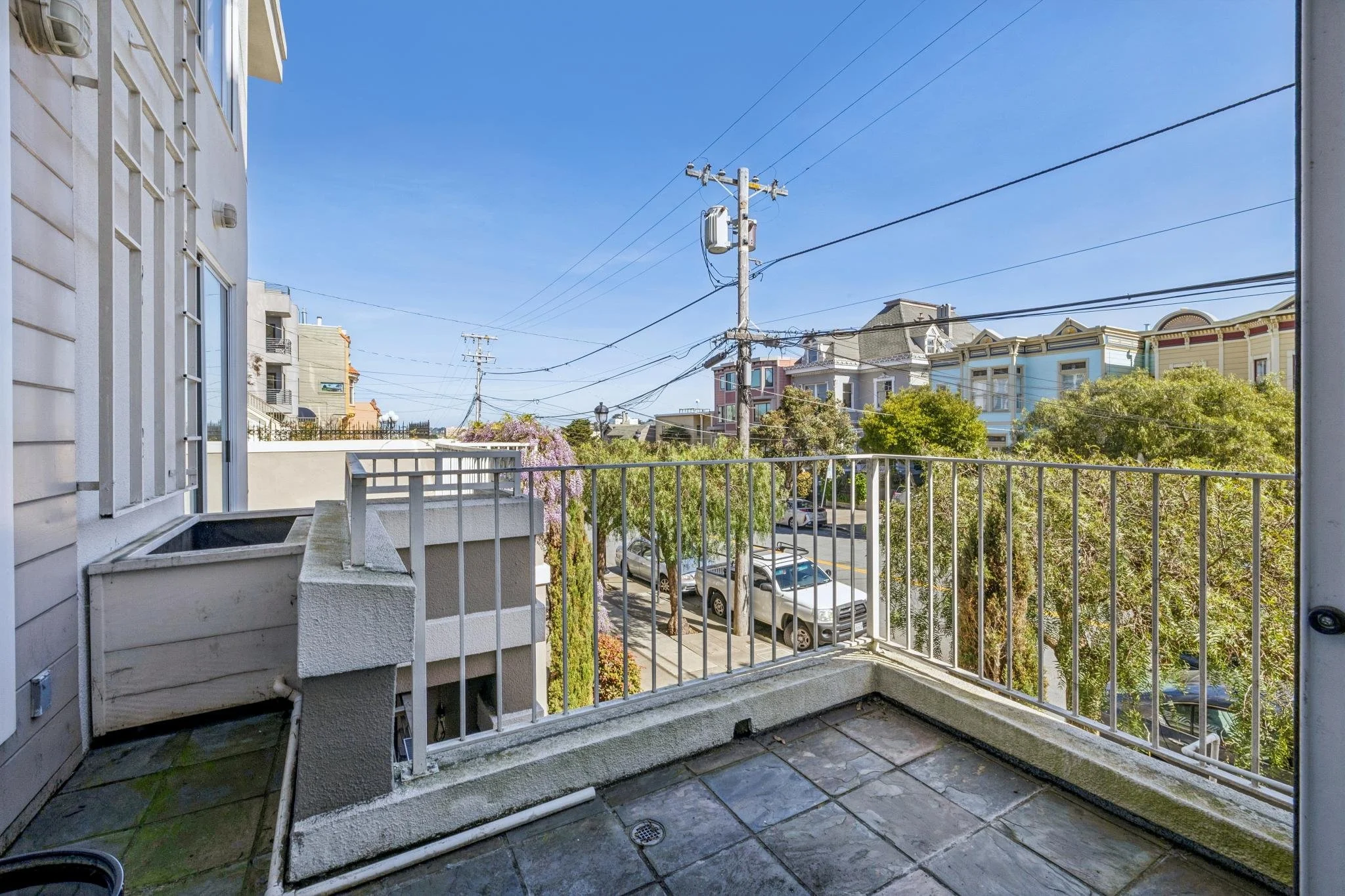 View from a small balcony showing a street with trees, parked cars, and Victorian-style houses under a blue sky.