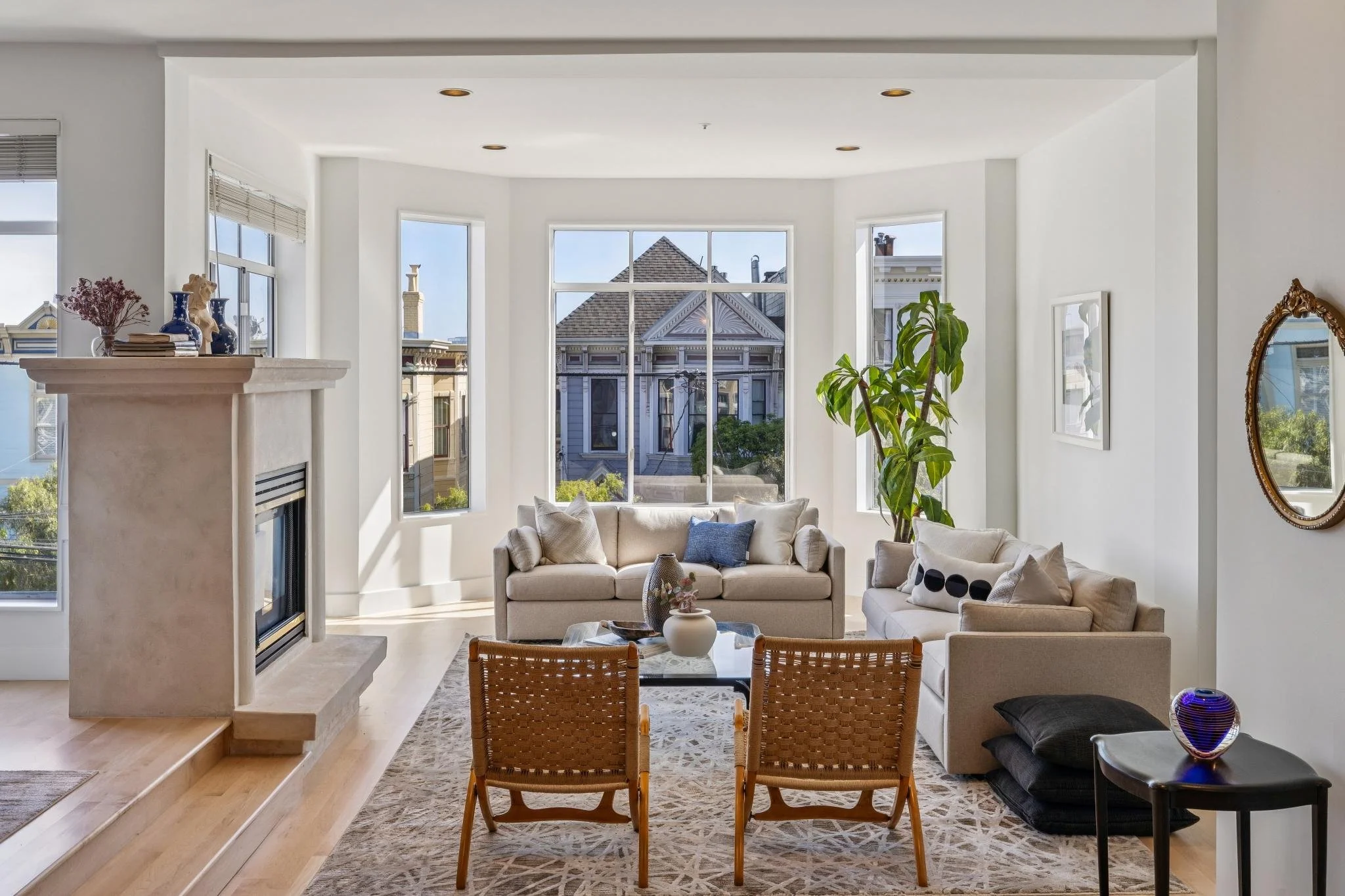 Bright living room with large bay windows, beige sofas, a glass coffee table, wooden chairs, a fireplace, and a tall green plant.