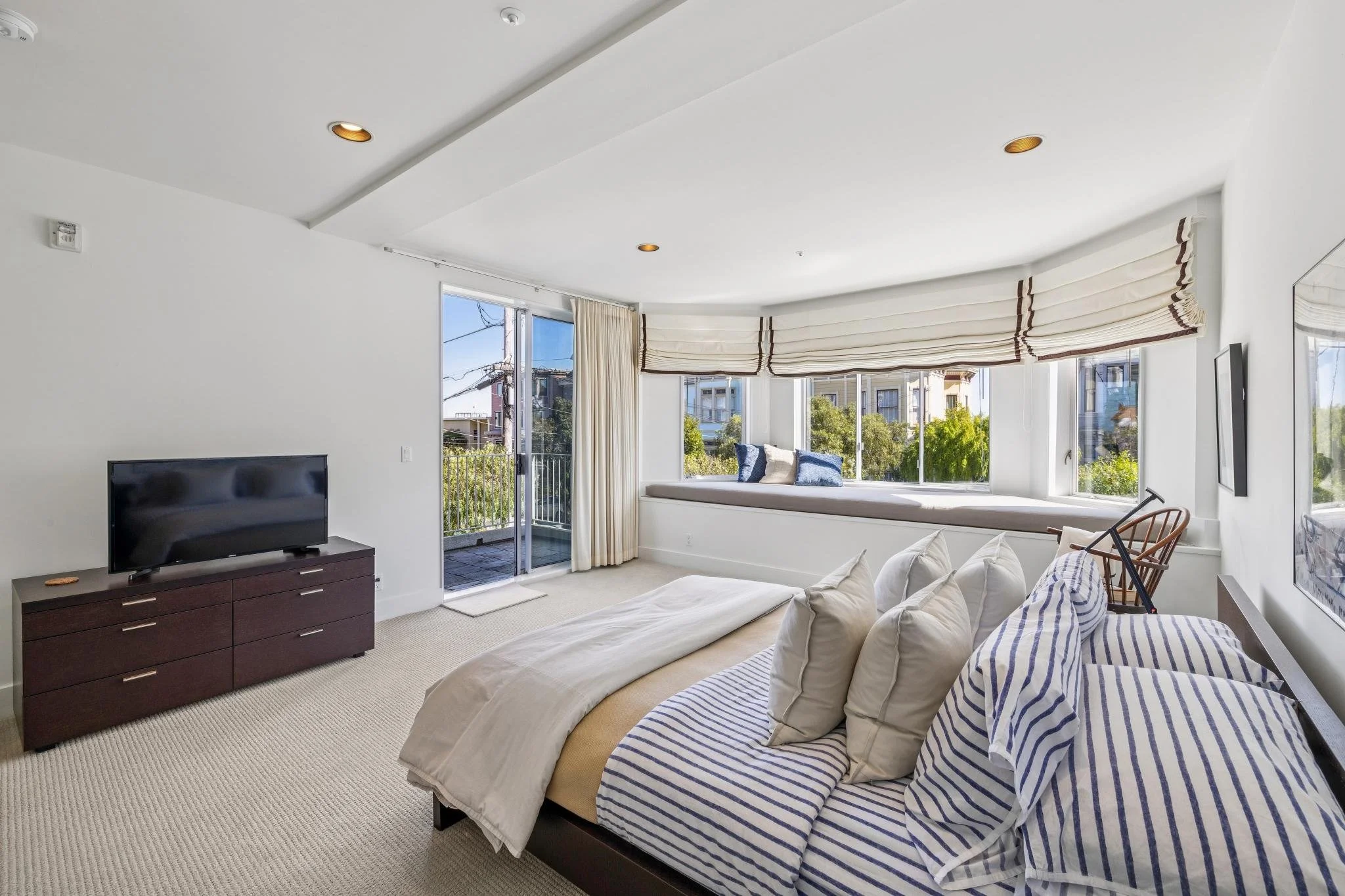 A bright bedroom with a bed, a window seat with cushions, and a sliding glass door leading to a balcony. A television on a dark wood dresser is also visible.