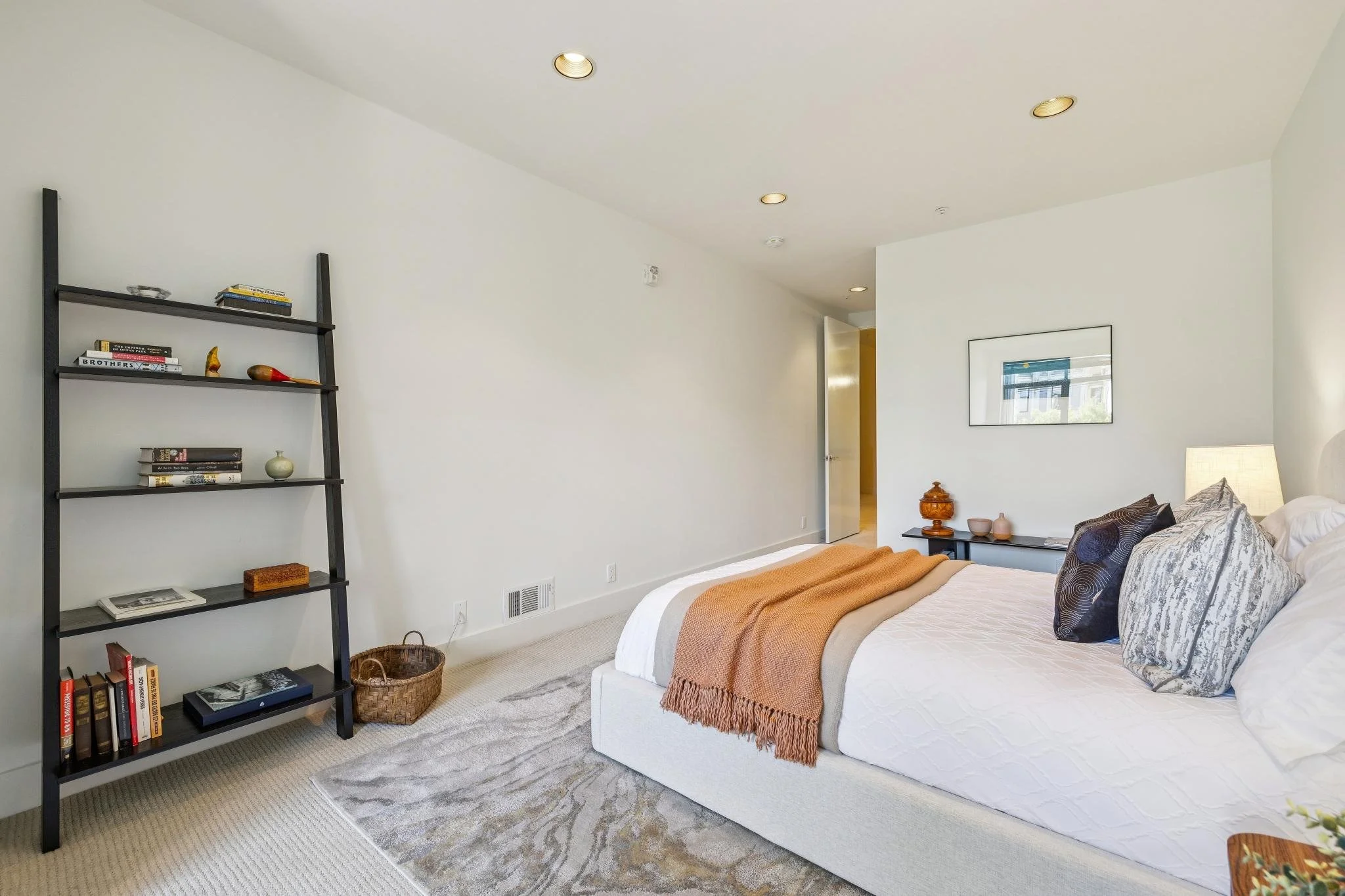 A modern bedroom with a white bed, colorful pillows, a burnt orange throw blanket, a black bookshelf on the left, and a window opposite the bed with a framed picture on the wall.