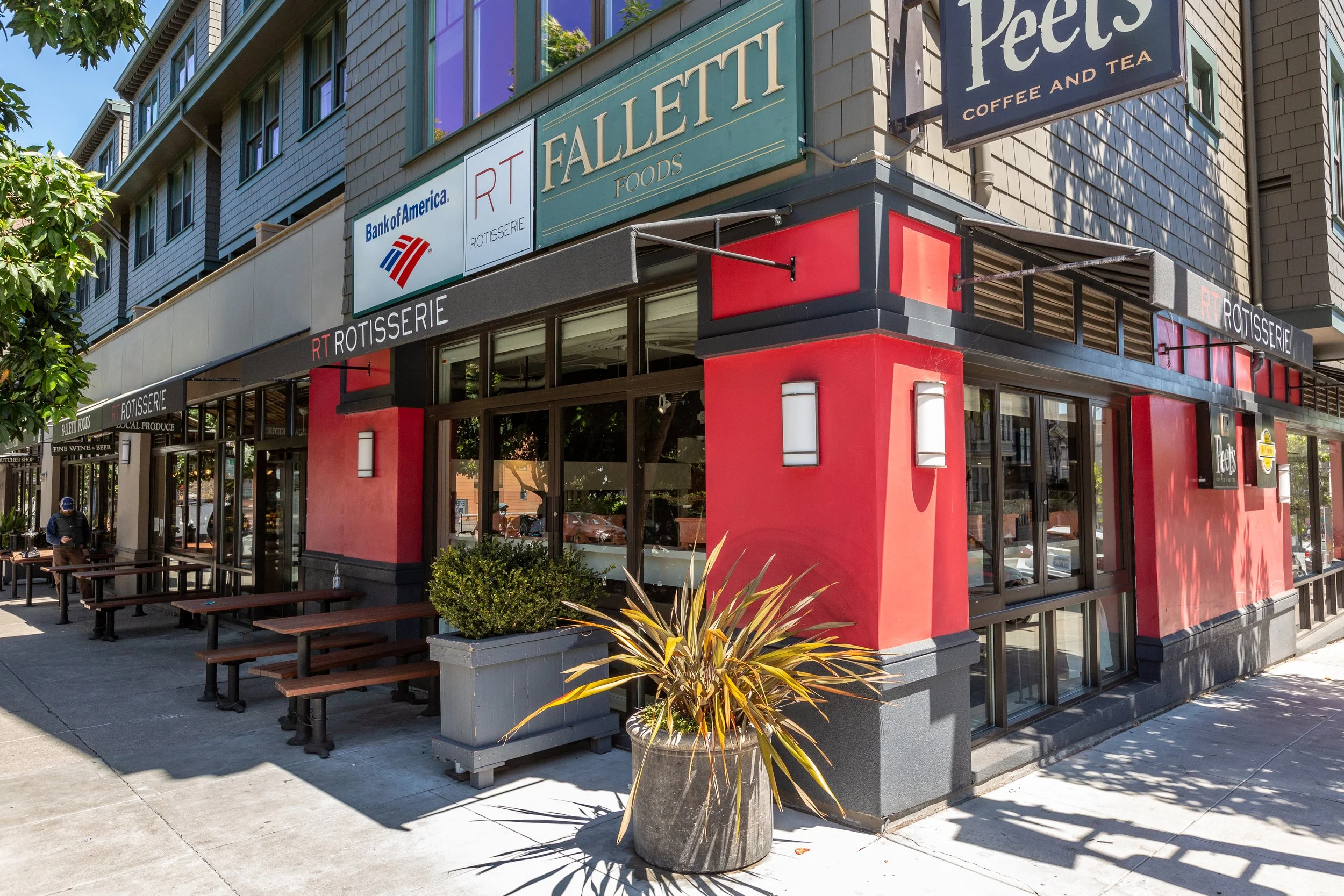 Exterior view of a restaurant named RT Rotisserie with red and black accents, outdoor seating, potted plants, and signage including 'Peet's Coffee and Tea' and 'Bank of America' on a sunny day.