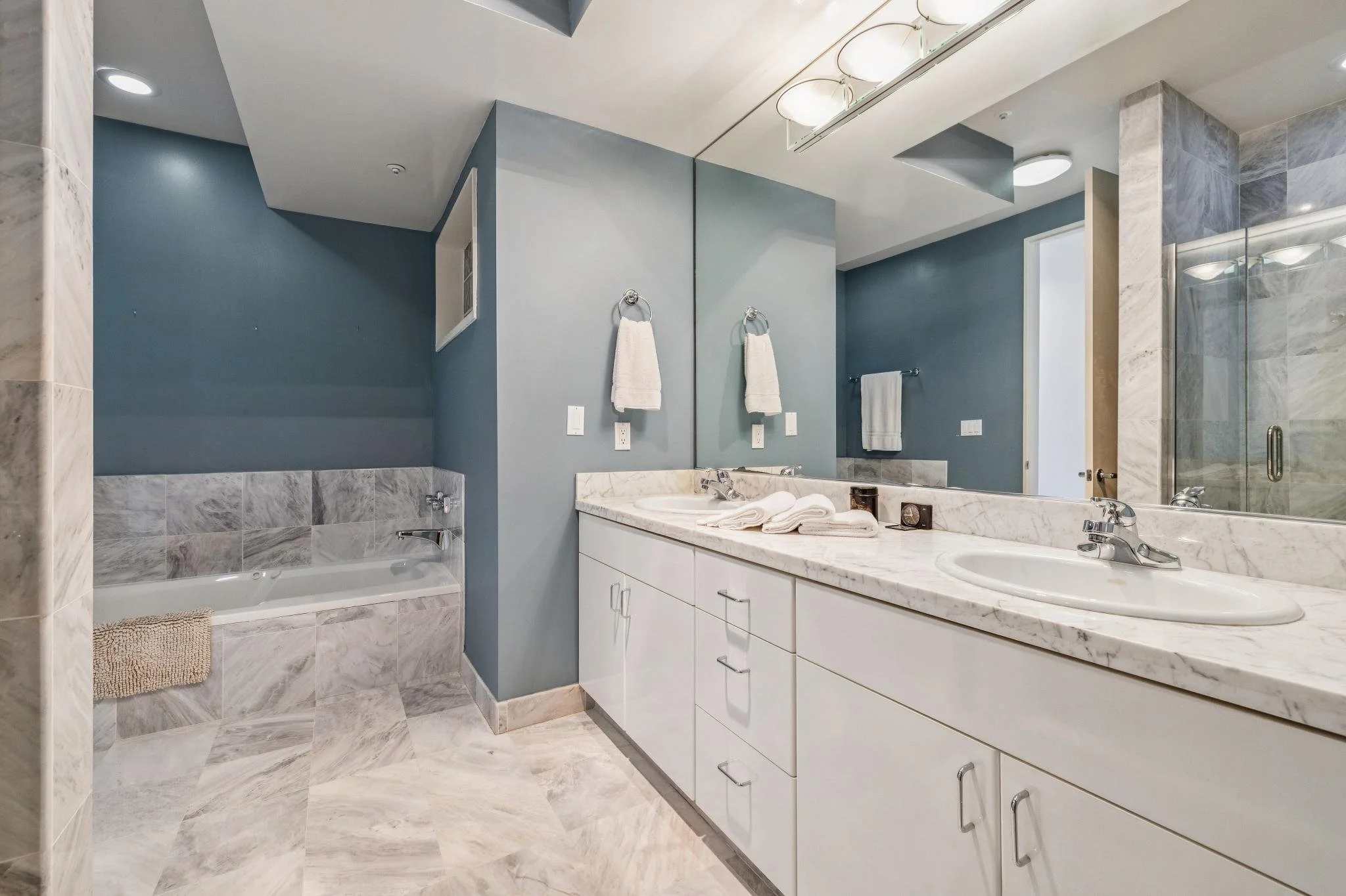 Modern bathroom with a double vanity, marble countertops, a large mirror, and a walk-in shower with glass doors. Towels are hung on the walls, and there is a bathtub with a marble surround in the corner.