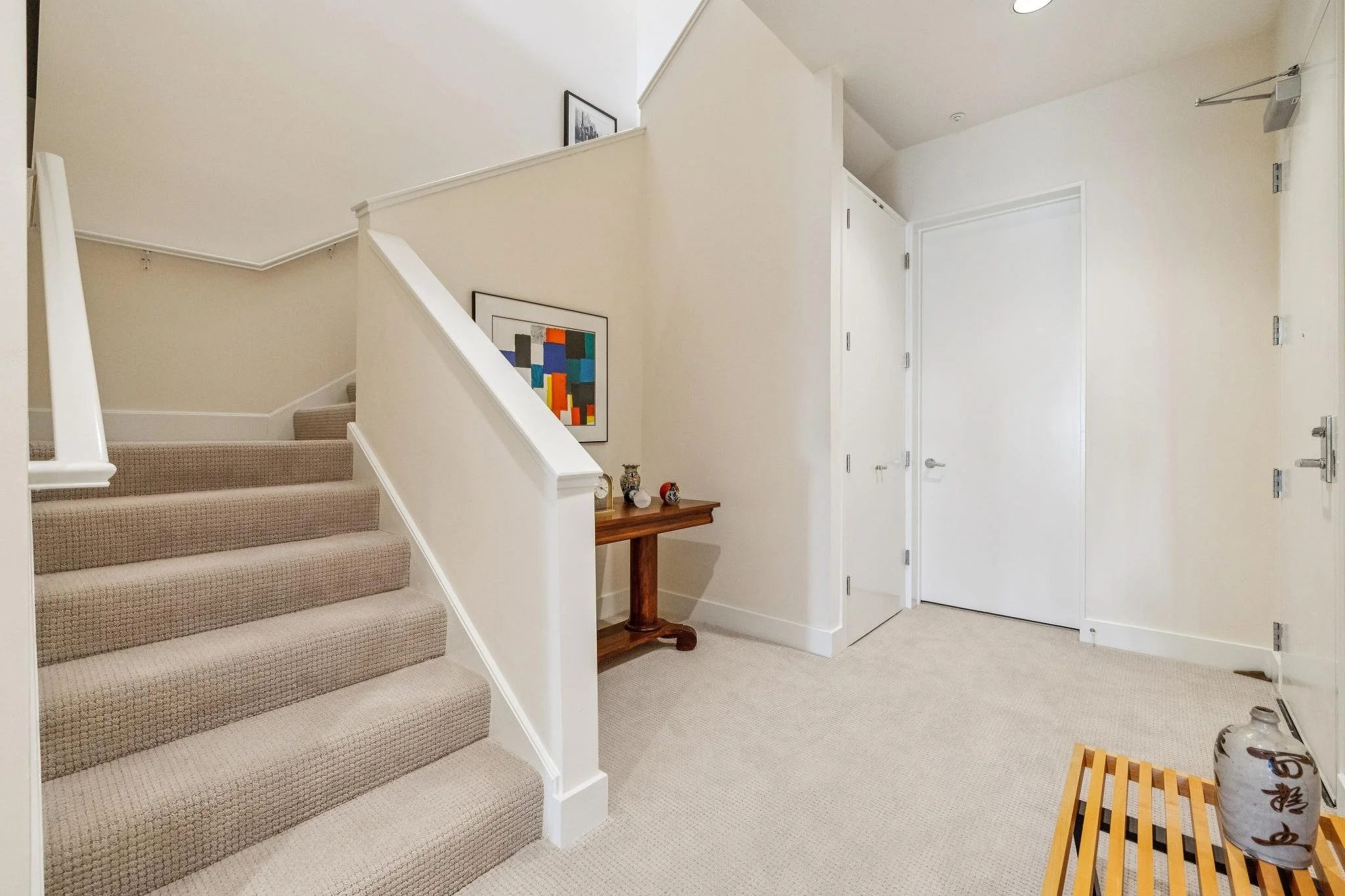 Interior view of a home entryway with beige carpeted stairs, white walls, a small wooden table with decorative items, and two white doors.