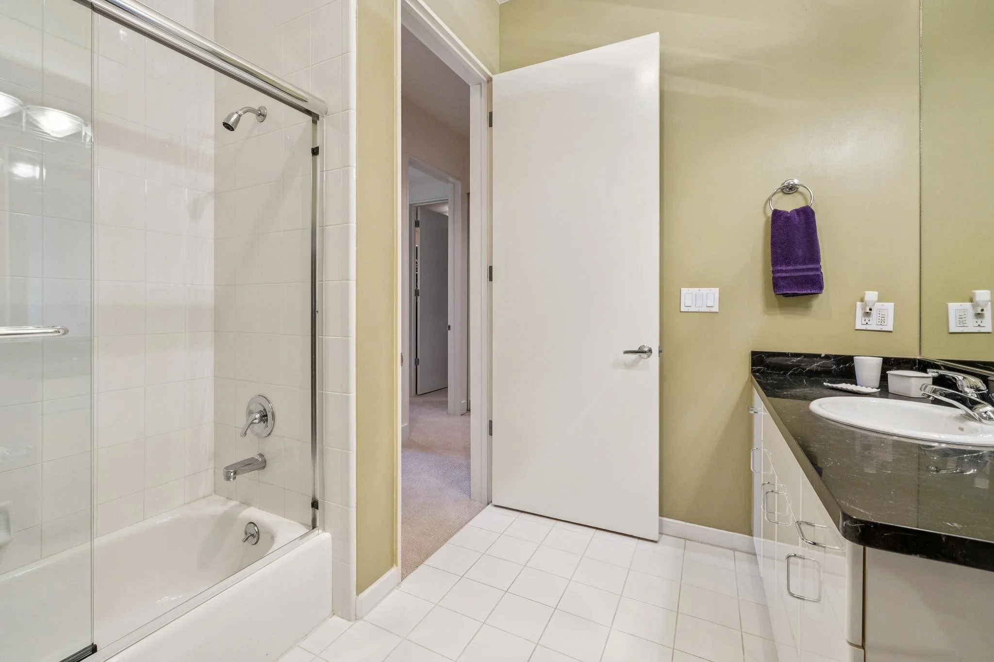Bathroom with a glass-enclosed shower, white tiled floor, beige walls, a white door, and a black countertop with a white sink. A purple towel hangs on a silver towel ring next to the mirror.
