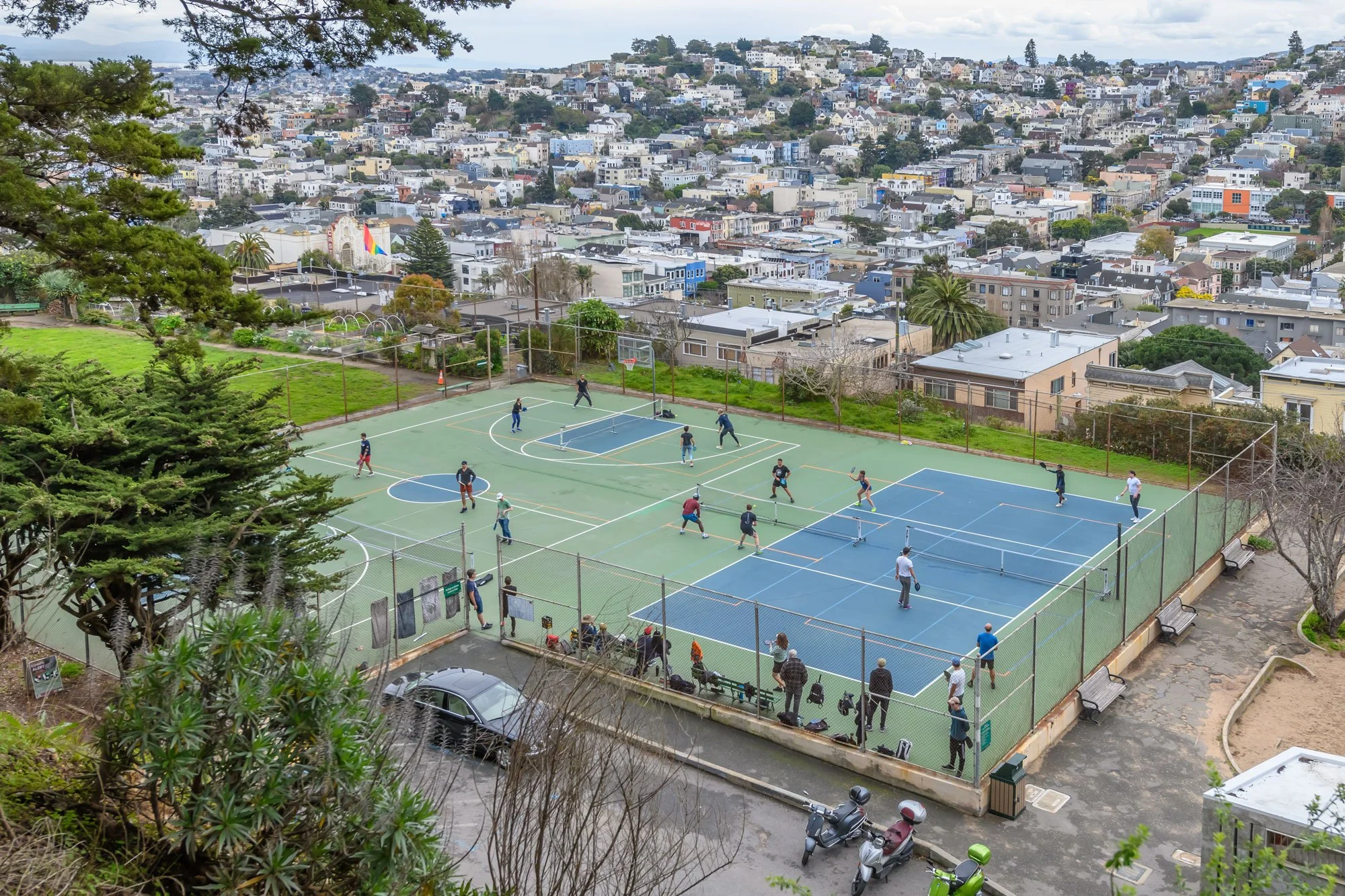 People playing tennis on multiple courts in an outdoor park, with a cityscape of densely packed buildings and homes in the background.