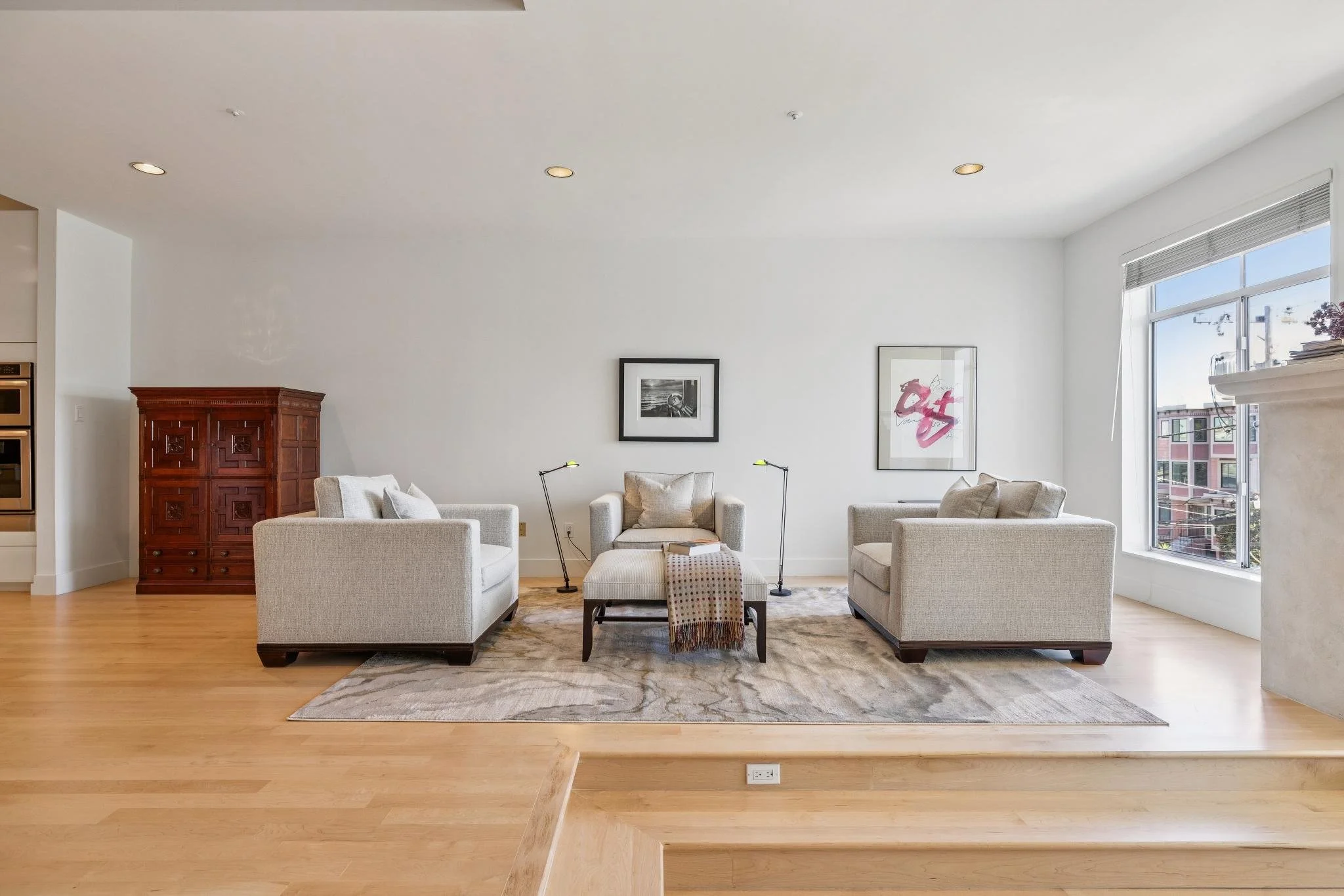 Living room with three white armchairs and a matching ottoman on a beige patterned rug, with two black floor lamps, white walls decorated with framed artwork, a big window, and a wooden floor.