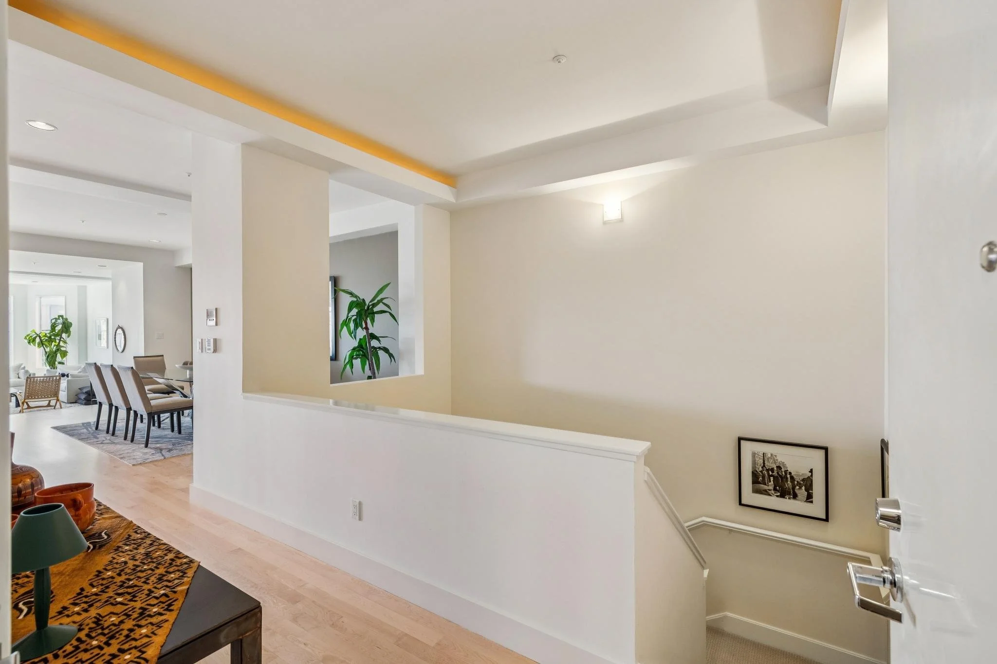 Interior view of an apartment with a staircase, framed black and white photograph on the wall, and living/dining area with chairs, plants, and natural lighting.