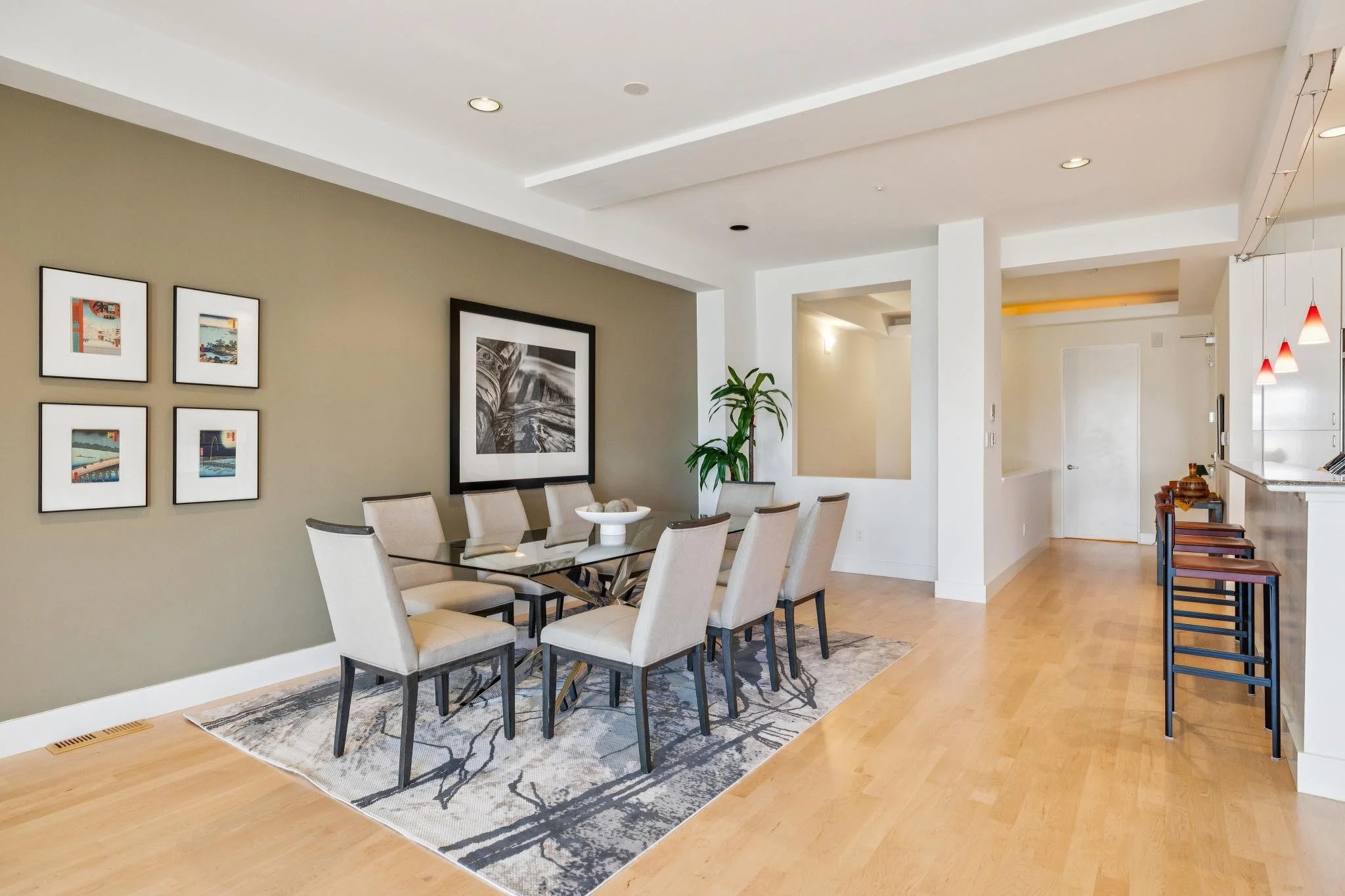 Dining area with a glass table, beige chairs, a patterned rug, and wall art, with a kitchen bar and stools visible in the background.