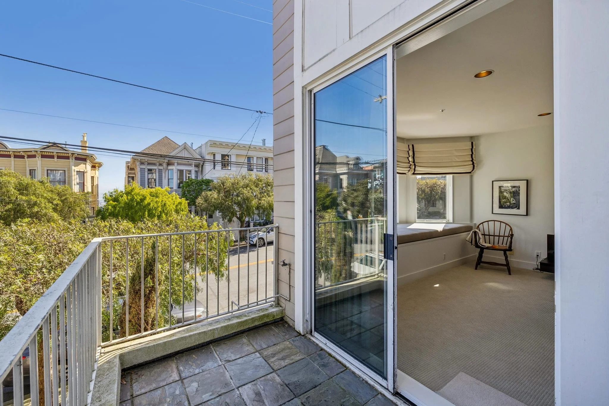Balcony connected to a living room with a sliding glass door, featuring city street view, trees, and residential buildings in the background.