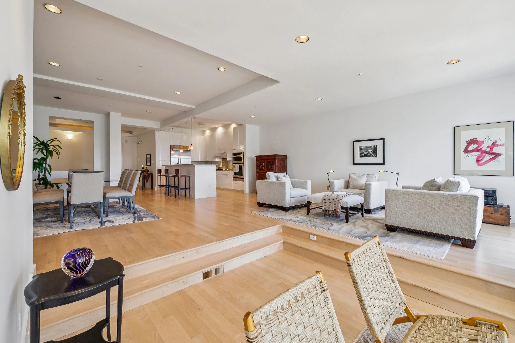 Bright open-concept living room and dining area with white walls, hardwood floors, and modern furniture, including a dining table with six chairs and a seating area with sofas and artwork on the wall.