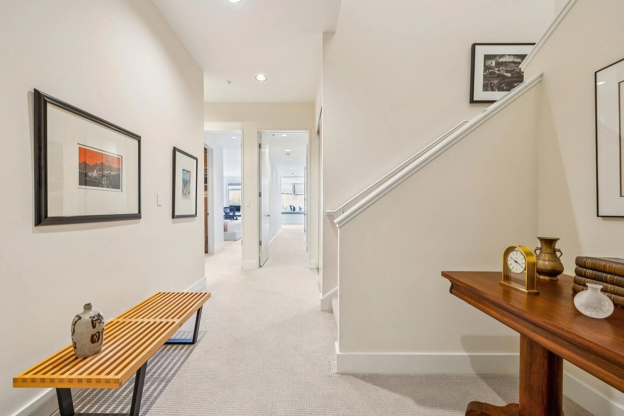An entryway with a hallway leading to a living room, decorated with wall art, a wooden bench on the left, and a dark wood console table on the right with decorative items, a clock, and books.