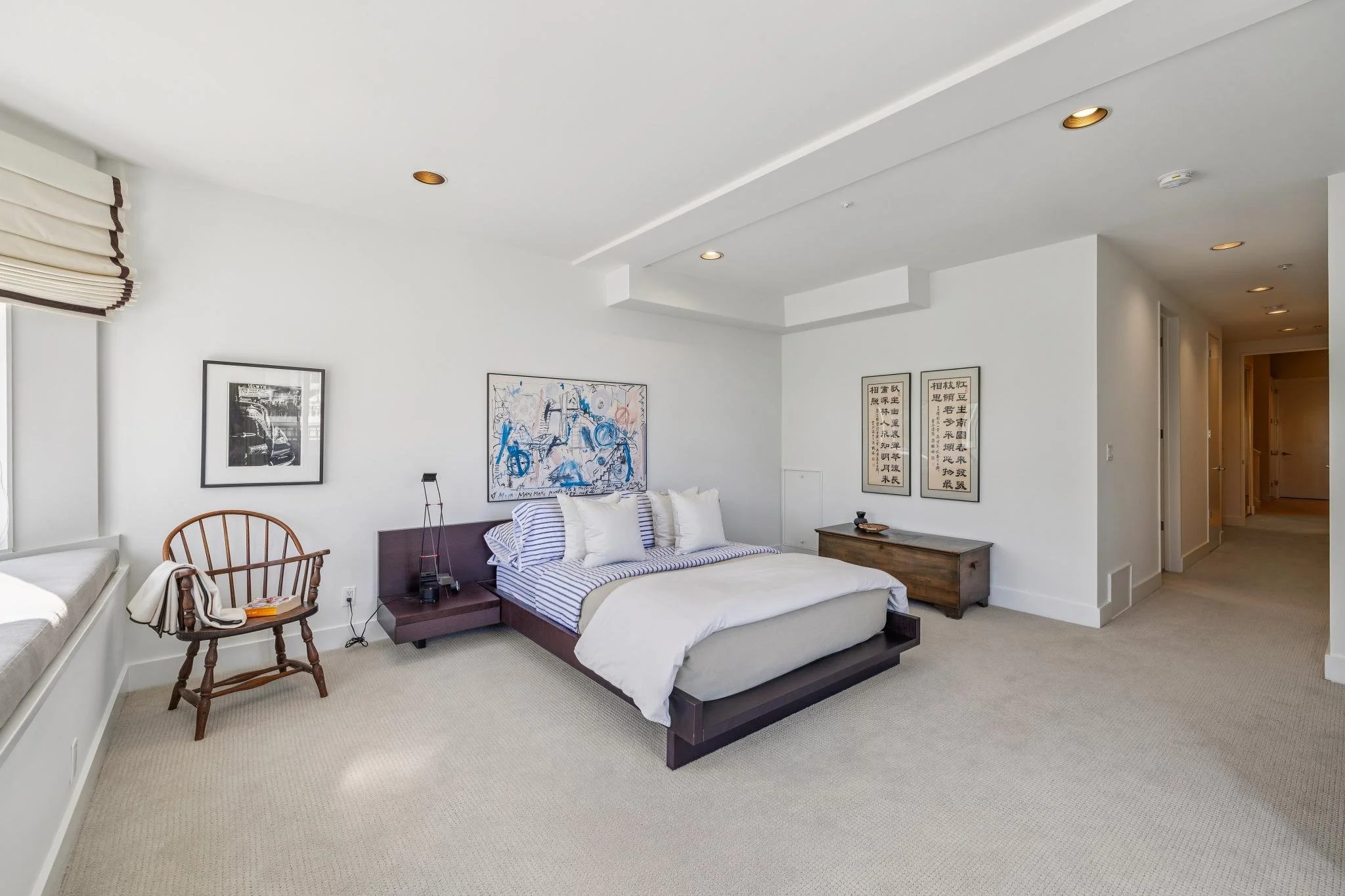 A minimalist bedroom with a white bed, striped bedding, wooden furniture, artwork, and a window seat with blinds.