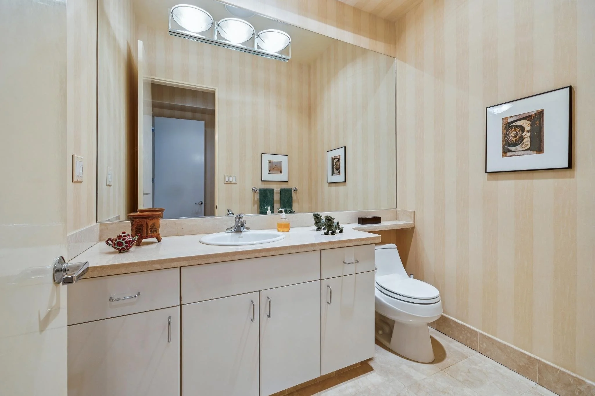 Bathroom with cream-colored walls, large mirror, beige countertop, white vanity with storage, small decorative items, and a toilet, with artwork on the walls.